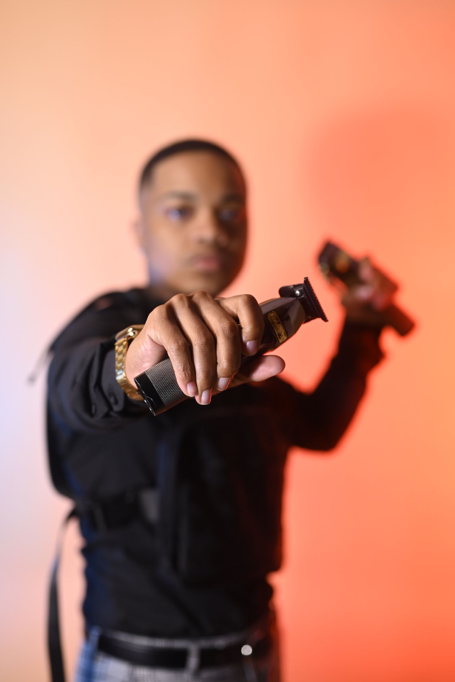 Barber in a black shirt and gold watch extends two hair clippers toward the camera against a vibrant orange studio backdrop.