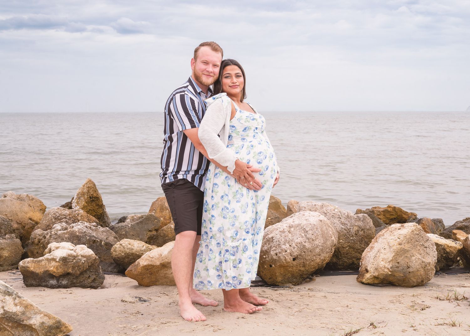 Pregnant couple posing on a rocky beach, woman in floral dress, man in striped shirt, ocean in background.