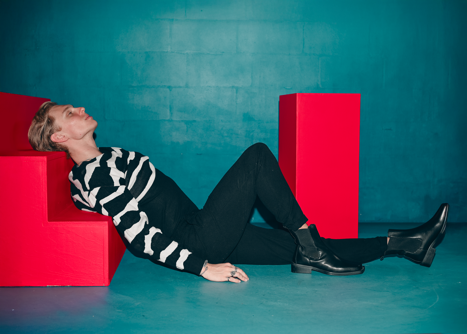 Artistic fashion shoot with model lying on floor against turquoise wall and red geometric shapes.