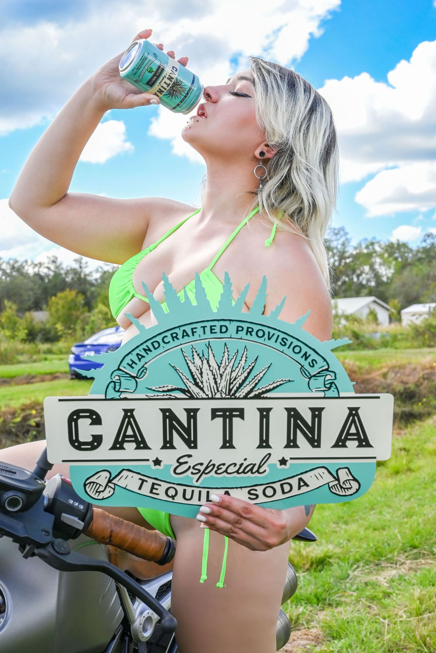 Woman in a green bikini sits on a motorcycle drinking a canned Cantina Especial tequila soda while holding the sign outdoors.