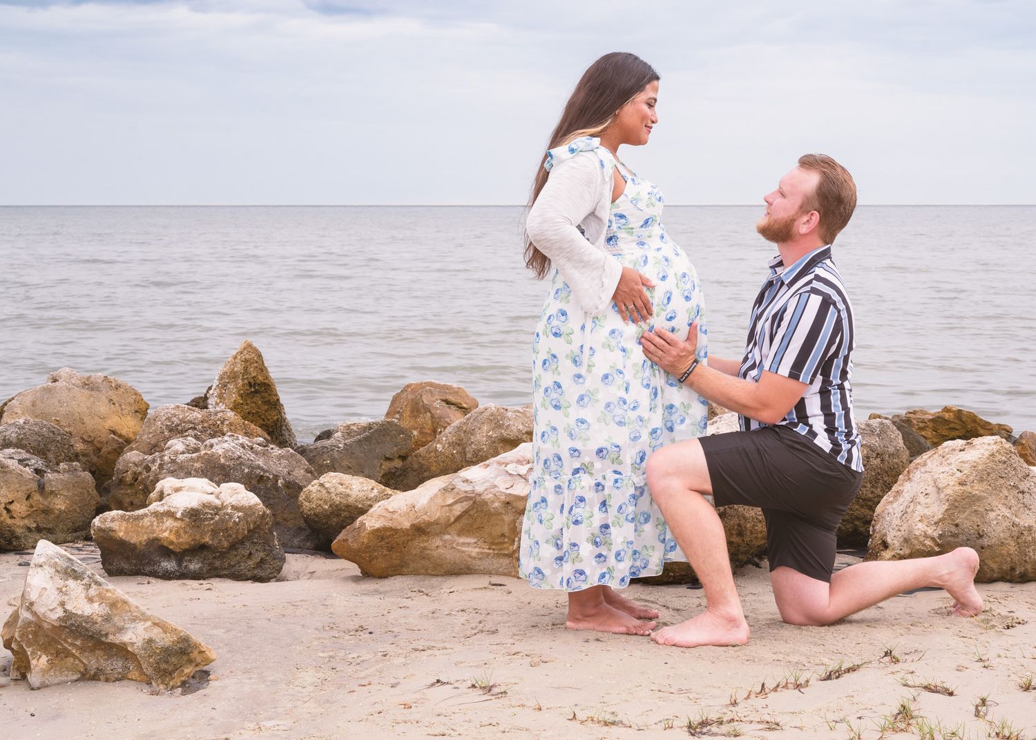 Pregnant woman in floral dress with partner kneeling on beach, touching baby bump near rocky shoreline.