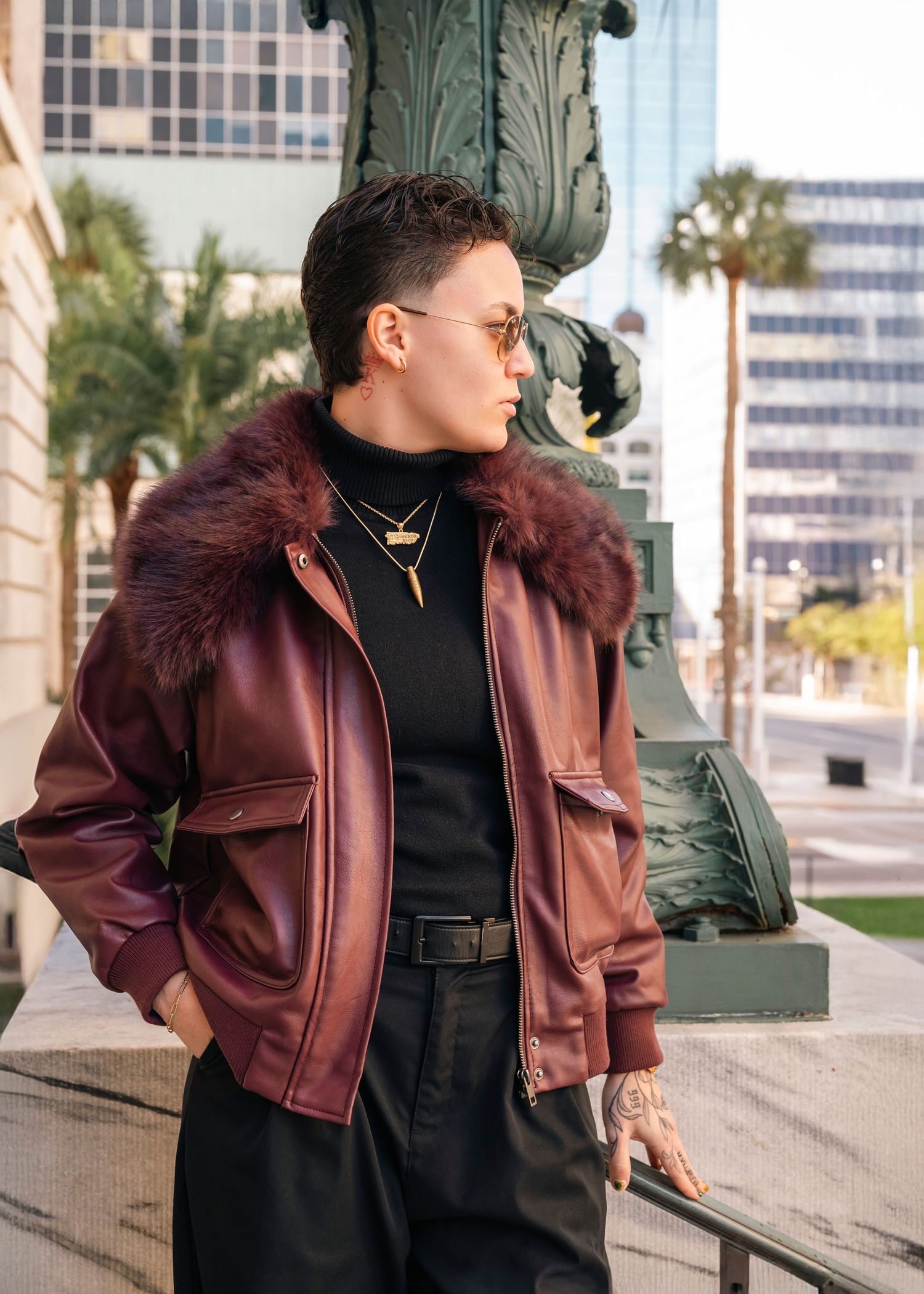 Person modeling burgundy leather jacket with fur collar against urban backdrop.