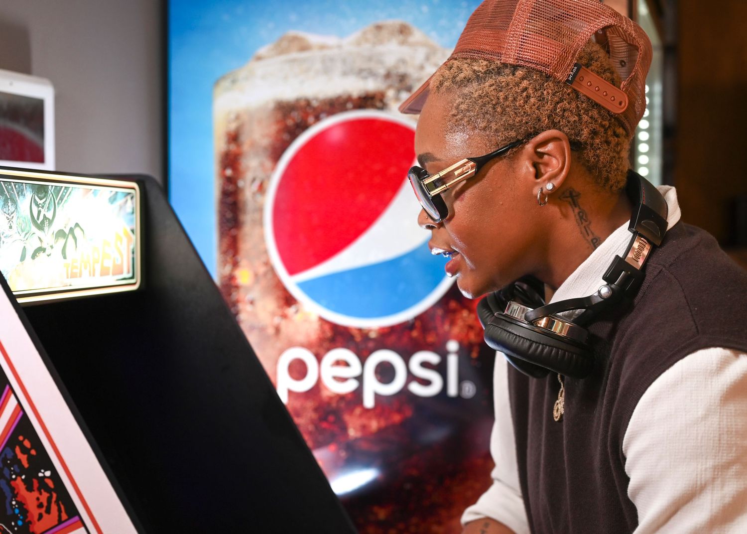 Person wearing headphones and a cap plays an arcade game with a Pepsi advertisement in the background.