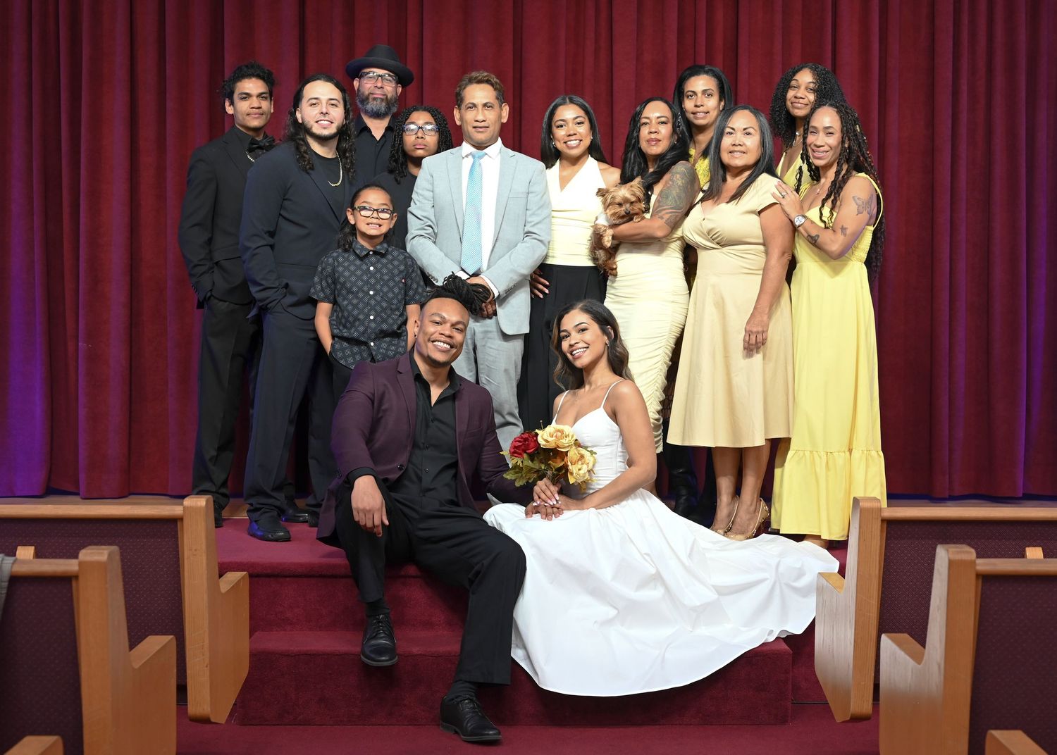Wedding party poses for a group photo in front of red curtains in a church setting.