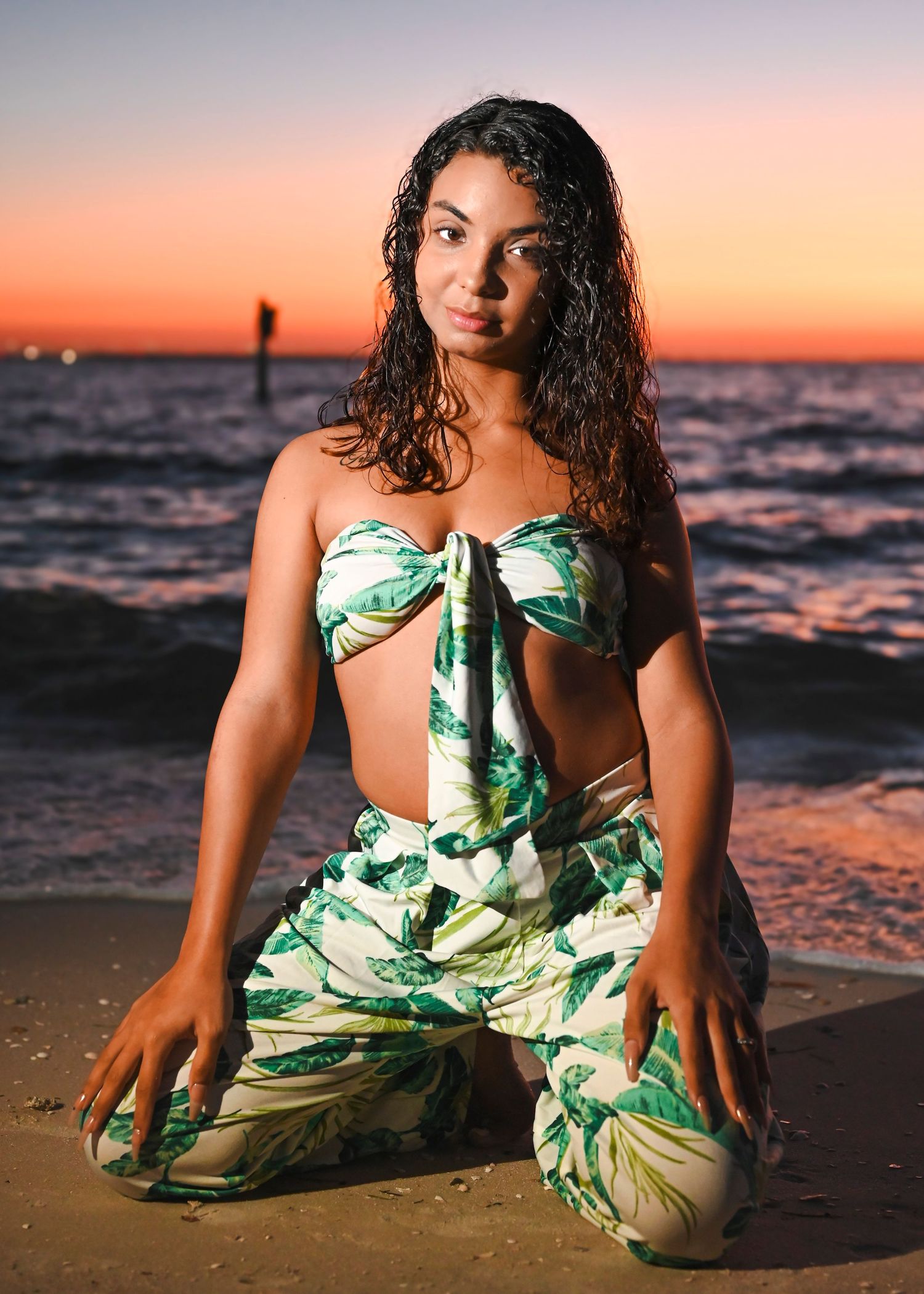 Young woman in tropical print outfit posing on sandy beach at sunset with ocean waves in background.