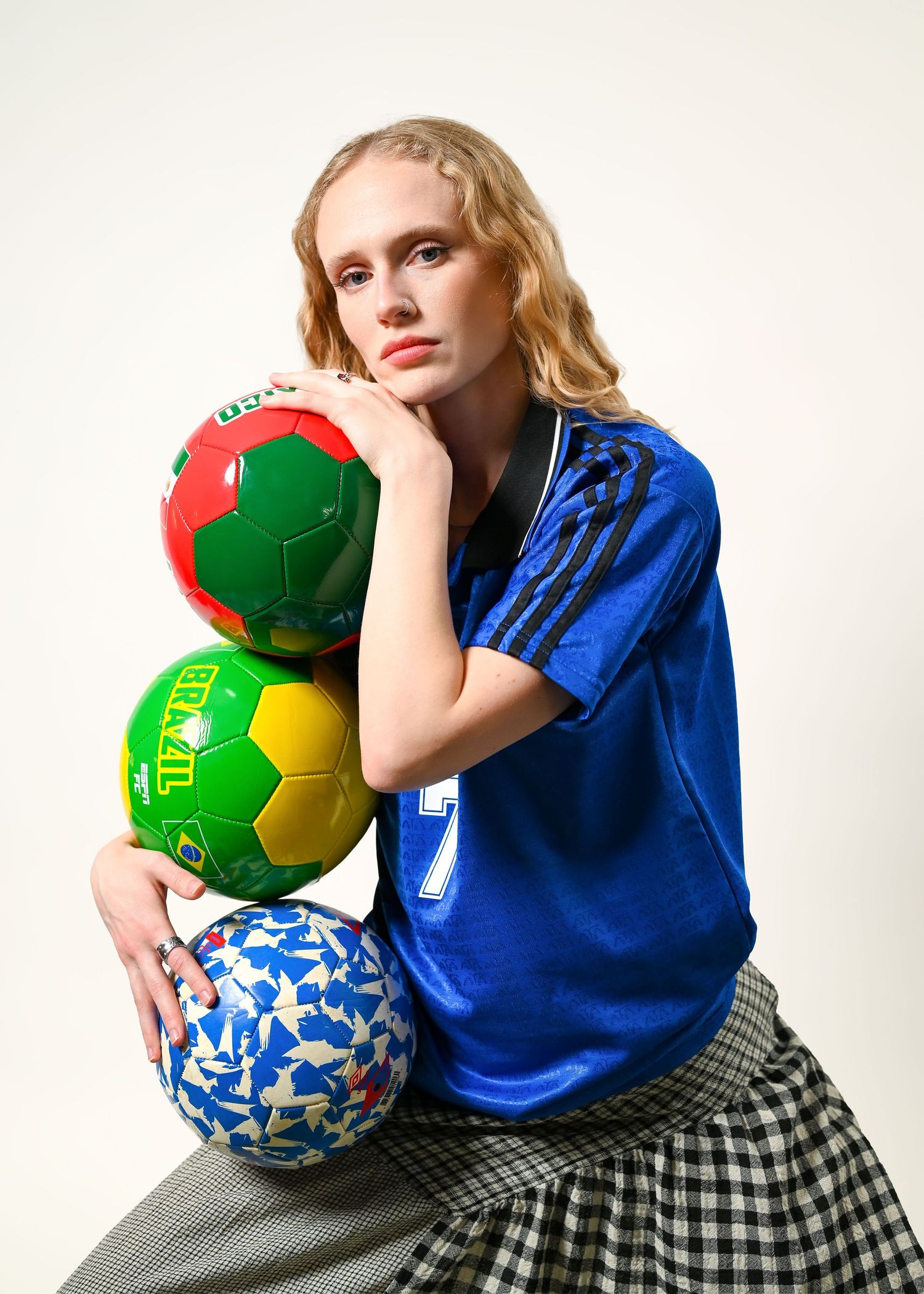 Woman in a blue soccer jersey and checkered skirt poses with three stacked colorful soccer balls, including a Brazil-themed ball.