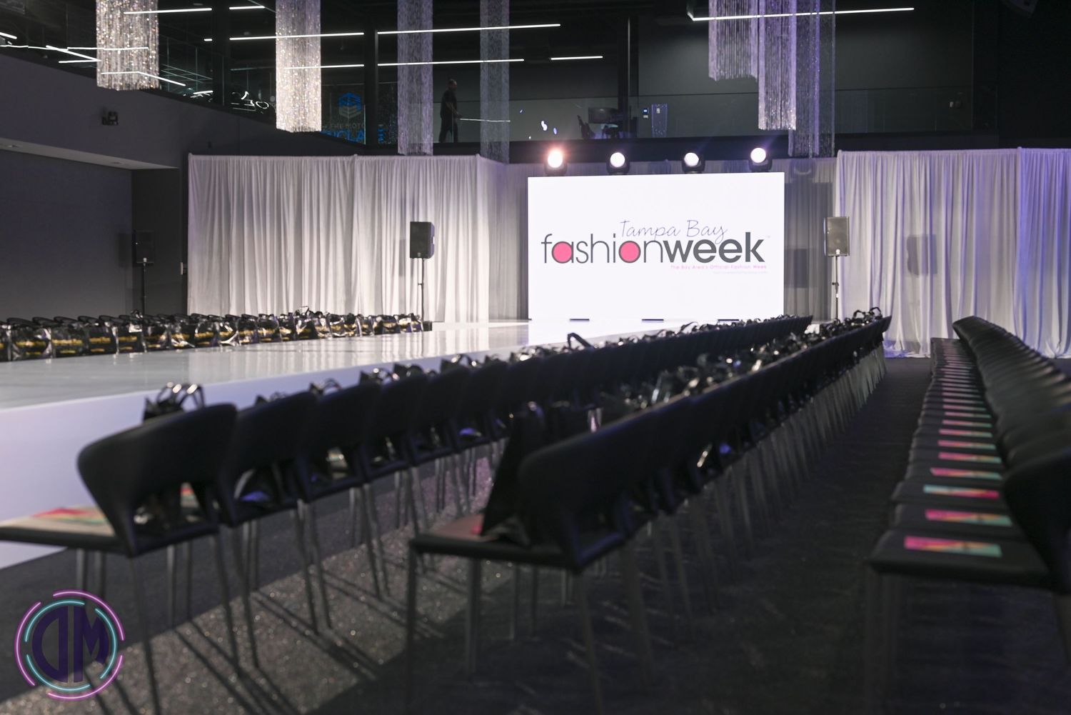 Dark fashion show venue with white runway and rows of empty black chairs.