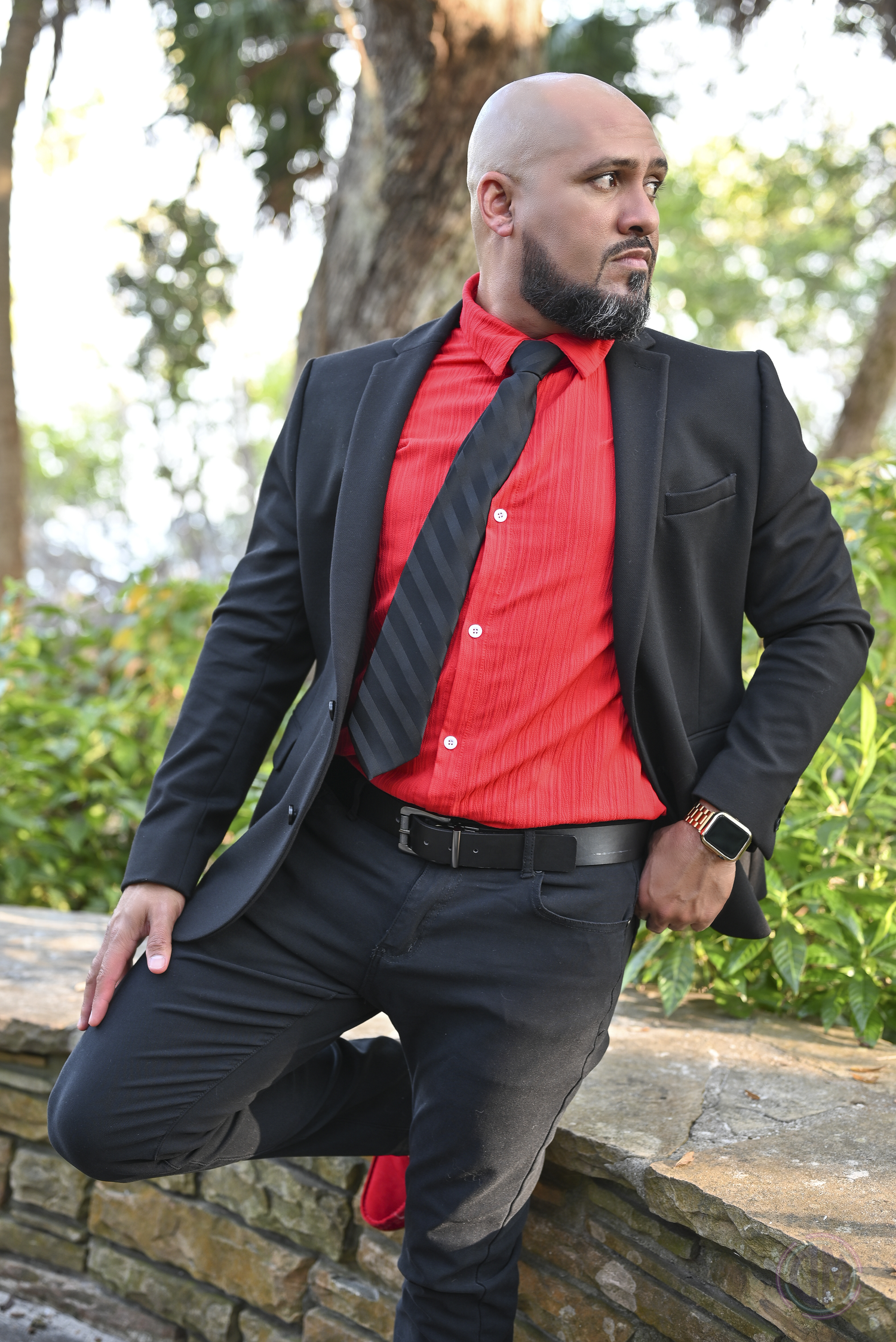Professional portrait in a black suit with red shirt and tie against outdoor garden setting.