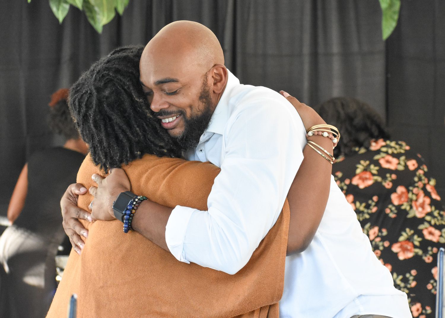 Two people share a warm, smiling embrace at an outdoor gathering while guests in floral clothing stand in the background.