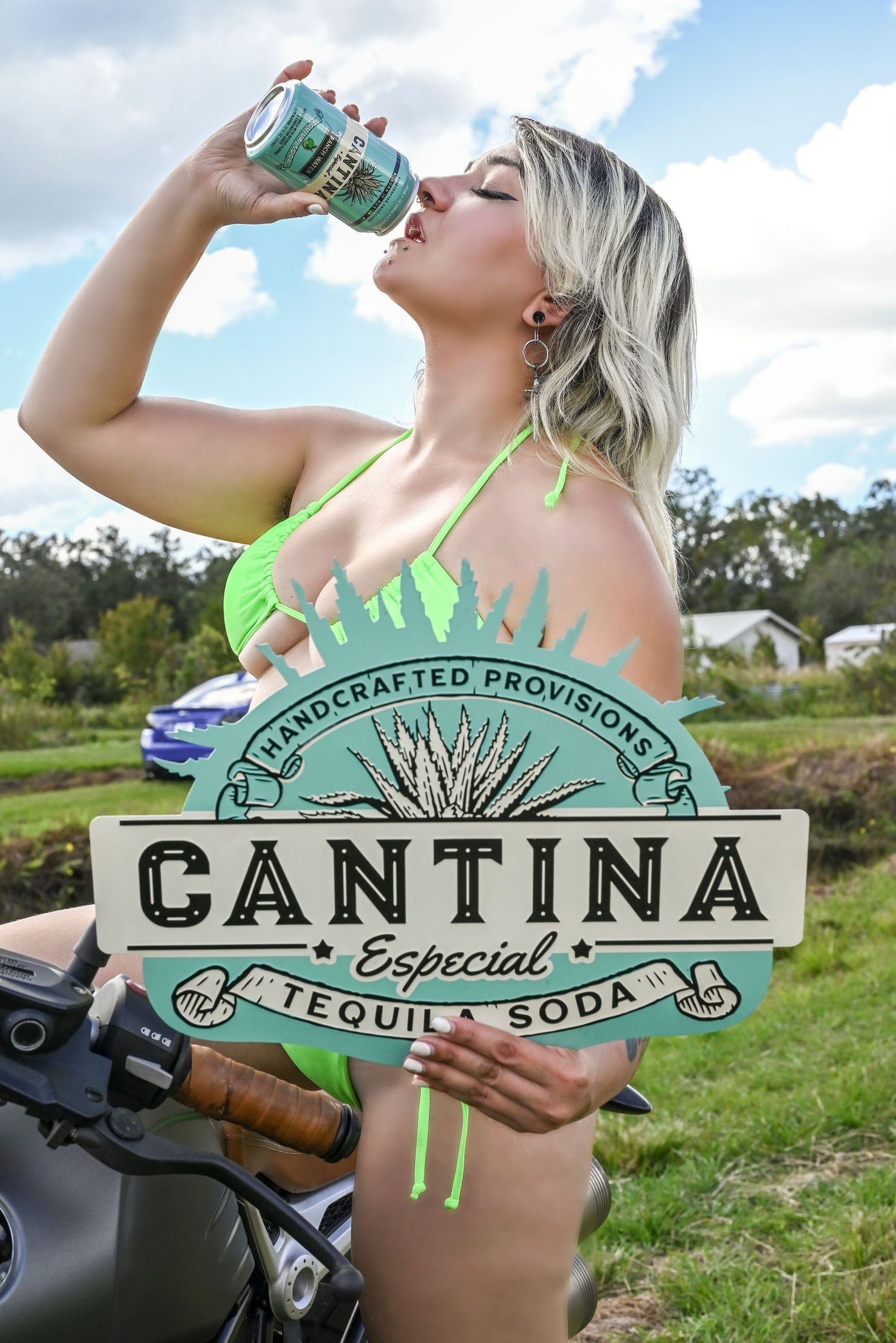 Woman in neon green swimwear drinking from a Cantina tequila soda can next to a motorcycle in an outdoor setting.