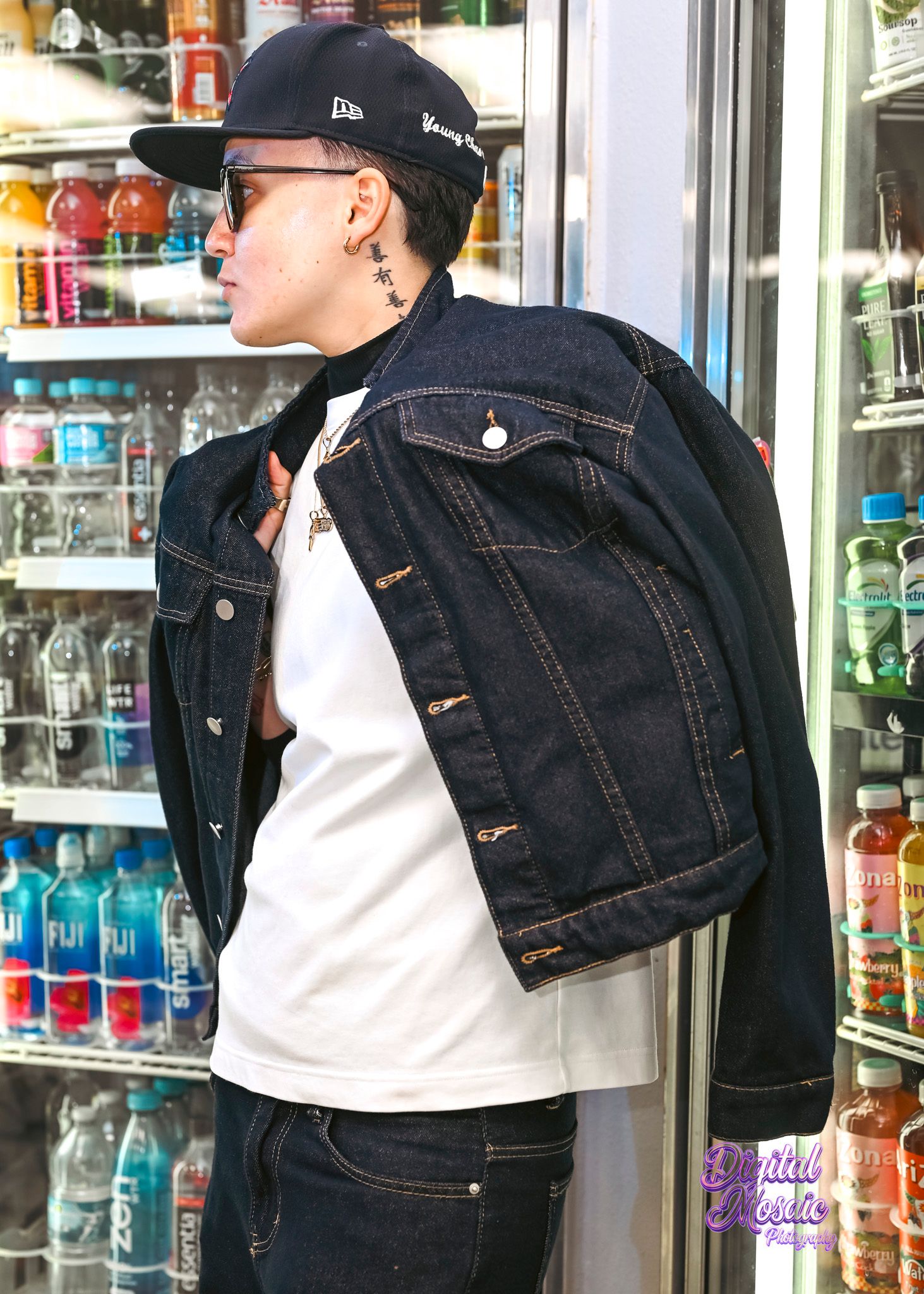 Person in a black cap, sunglasses, dark denim jacket and white tee stands beside a convenience store cooler of colorful bottled drinks.