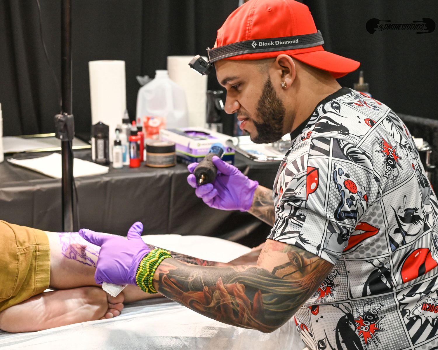 Tattoo artist wearing gloves works on client at convention booth.