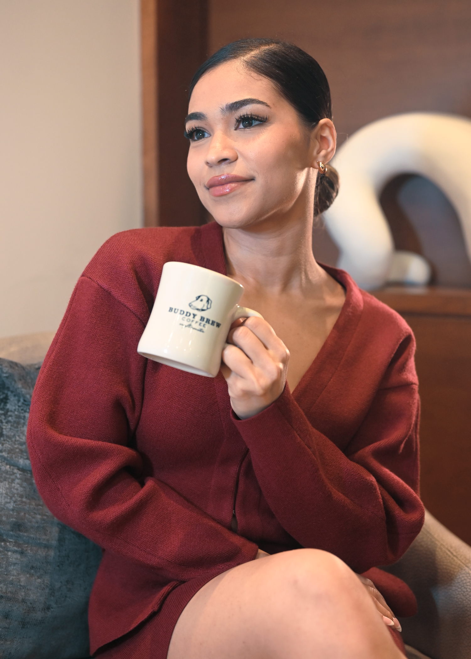 Cozy indoor scene with someone in burgundy sweater holding coffee cup on grey couch.