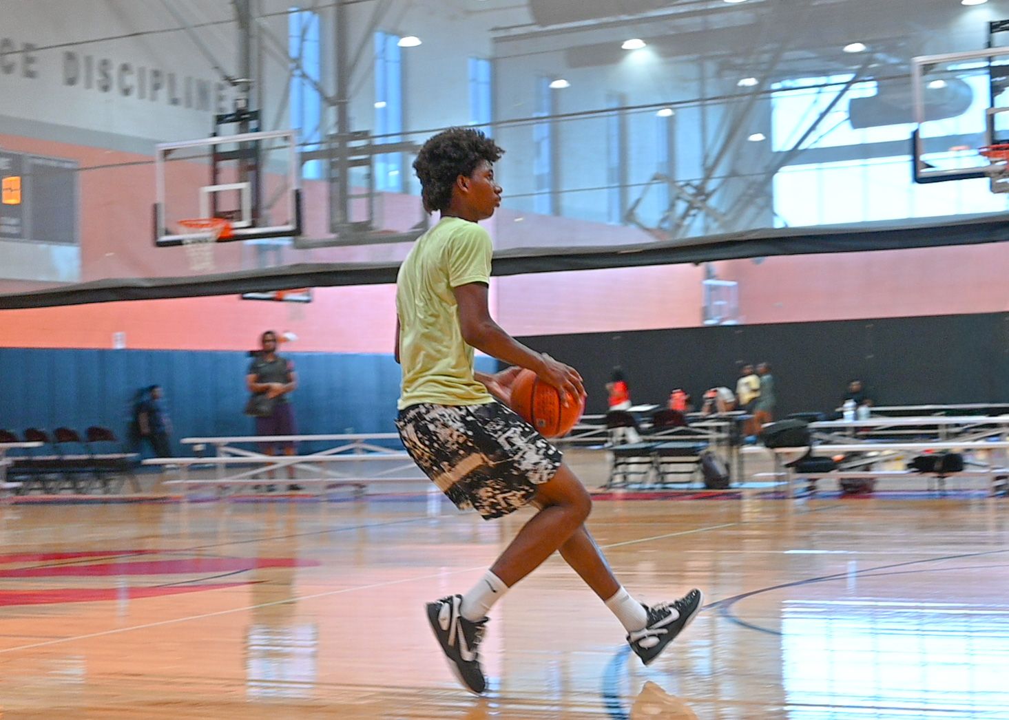 Young basketball player in a yellow shirt and patterned shorts dribbles a basketball across an indoor gym court during practice.
