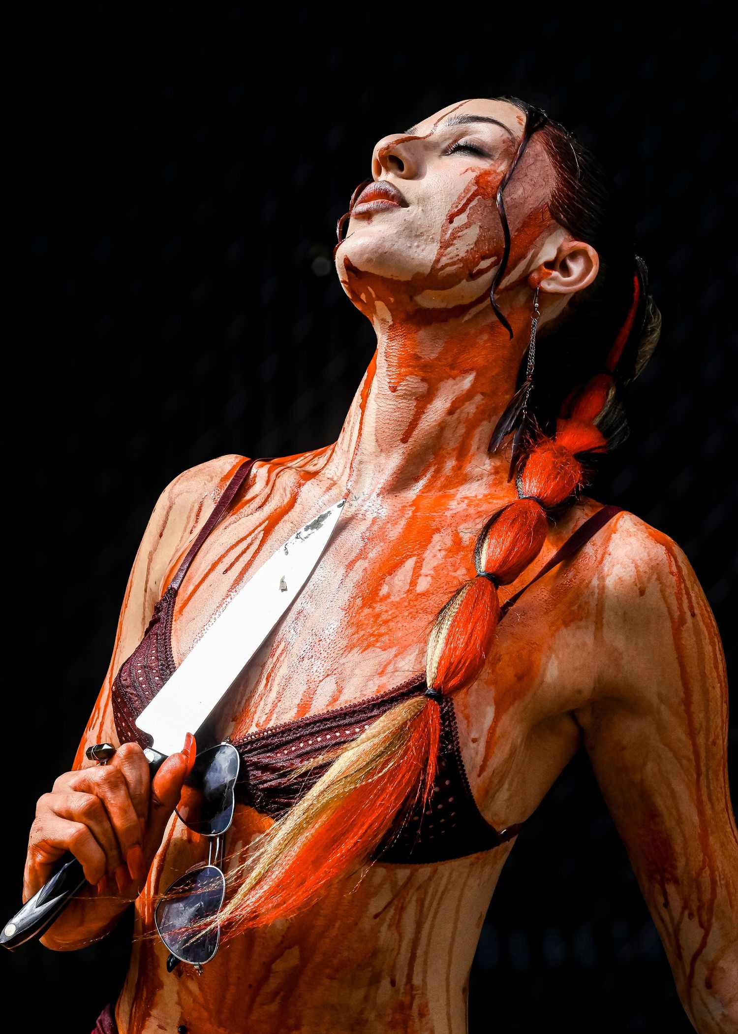Woman with an orange braid and head tilted back, covered in red fake blood, holding a large knife against a dark background.
