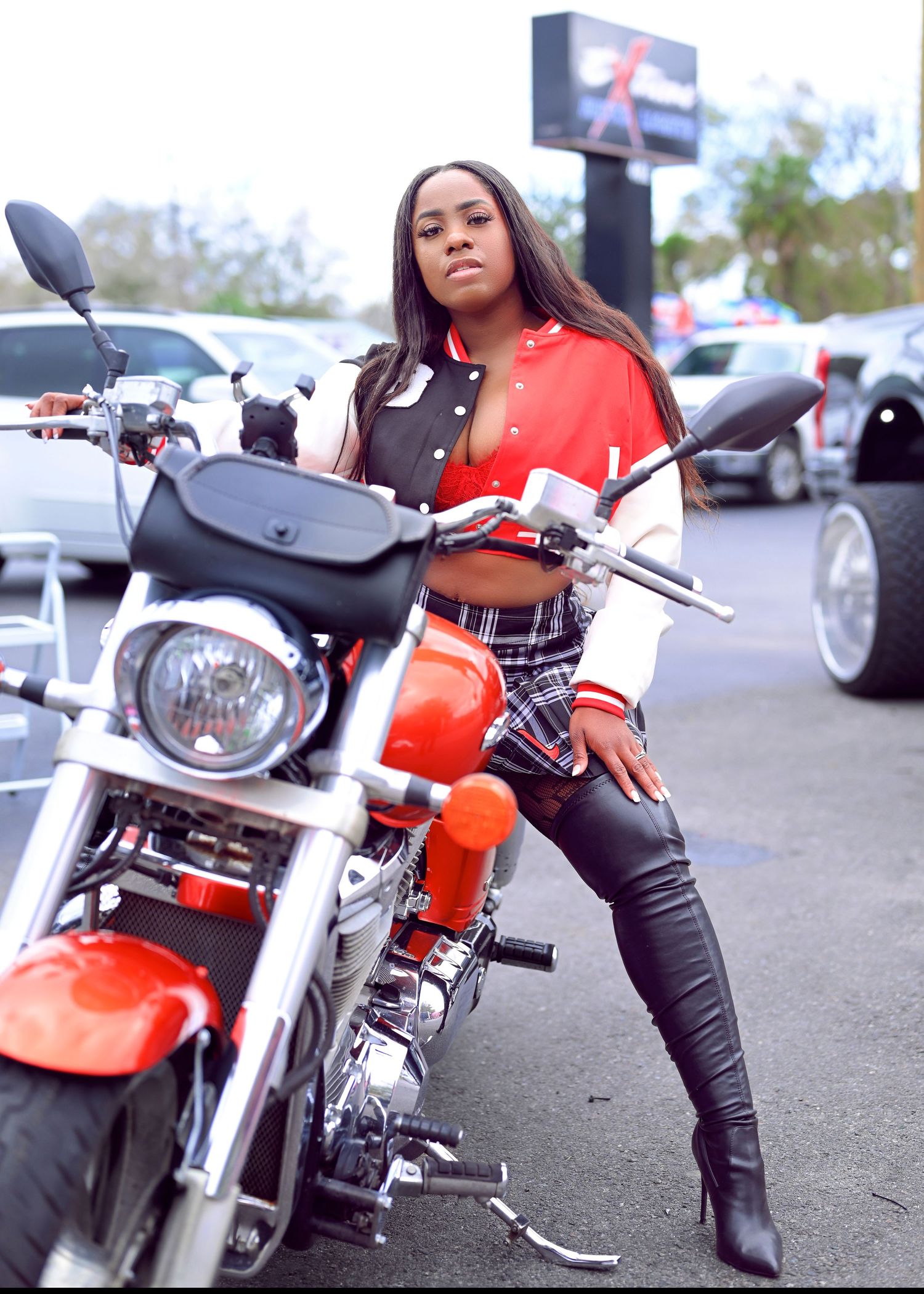 Model in black outfit poses with red motorcycle in parking lot.