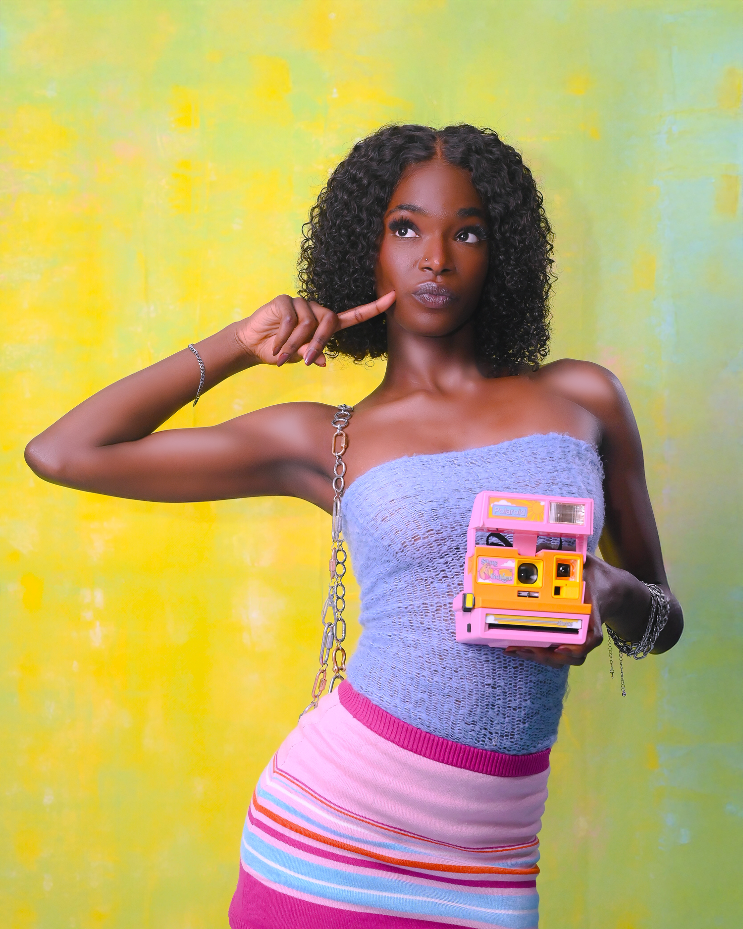 Vibrant fashion portrait with curly hair holding pink camera against yellow-green background.
