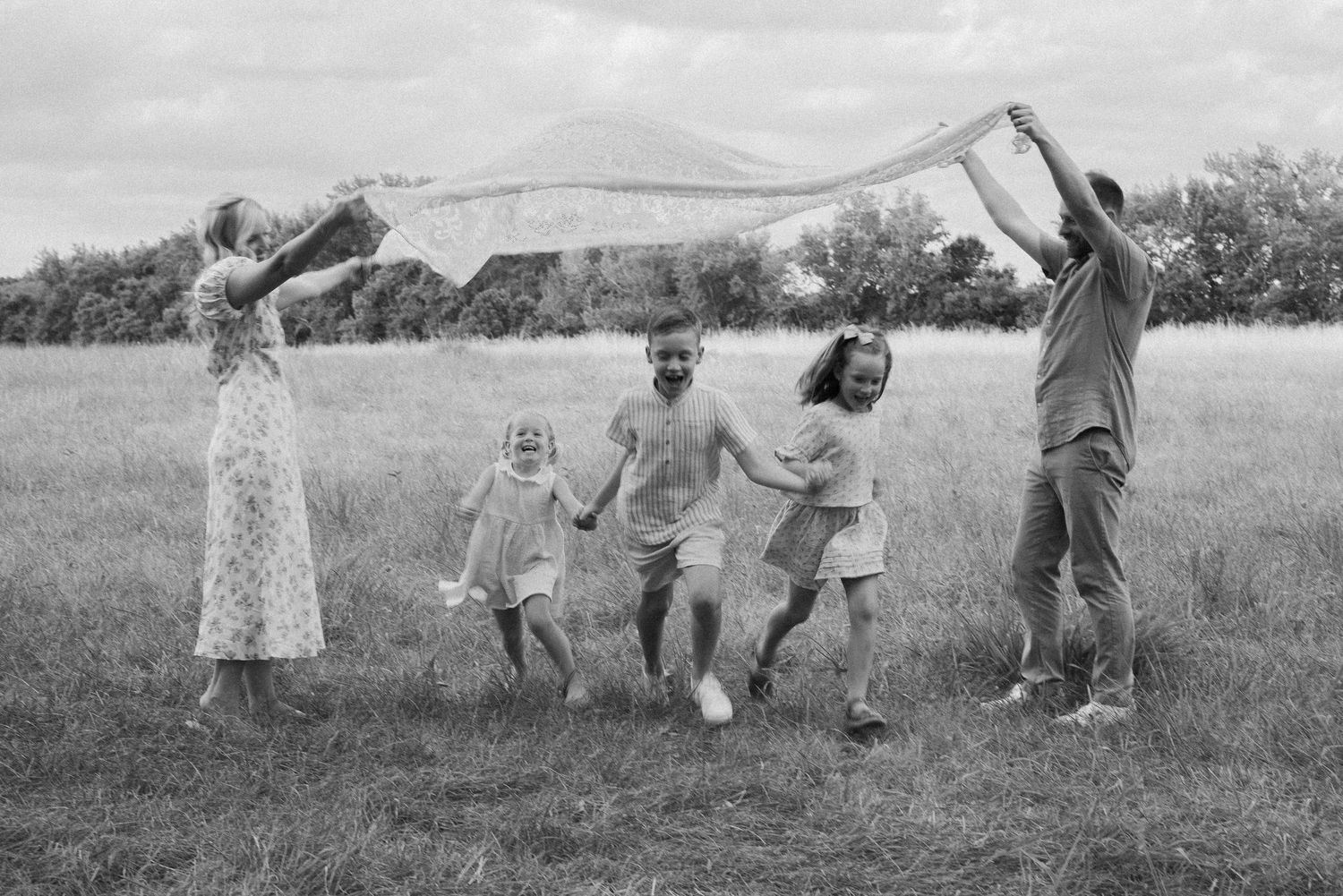 mom and dad holding blanket. three children running underneath. smiling. laughing. toddlers. brother, sister. black and white photo in field