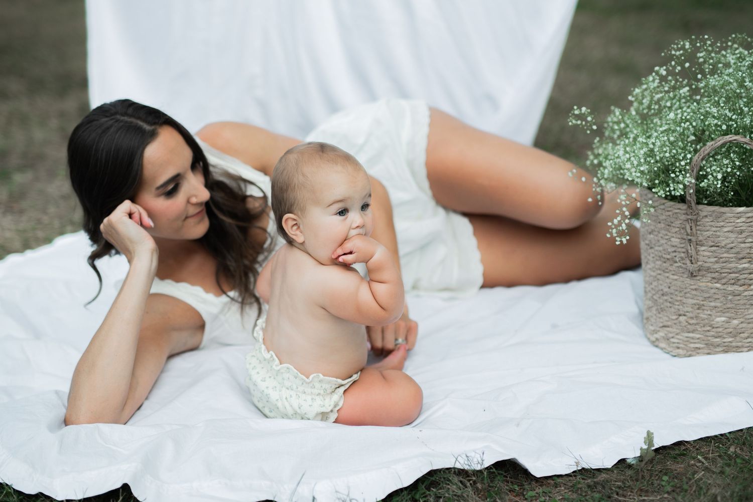 mom and baby outside laying on white sheet with clothesline hugging, baby's breath in basket