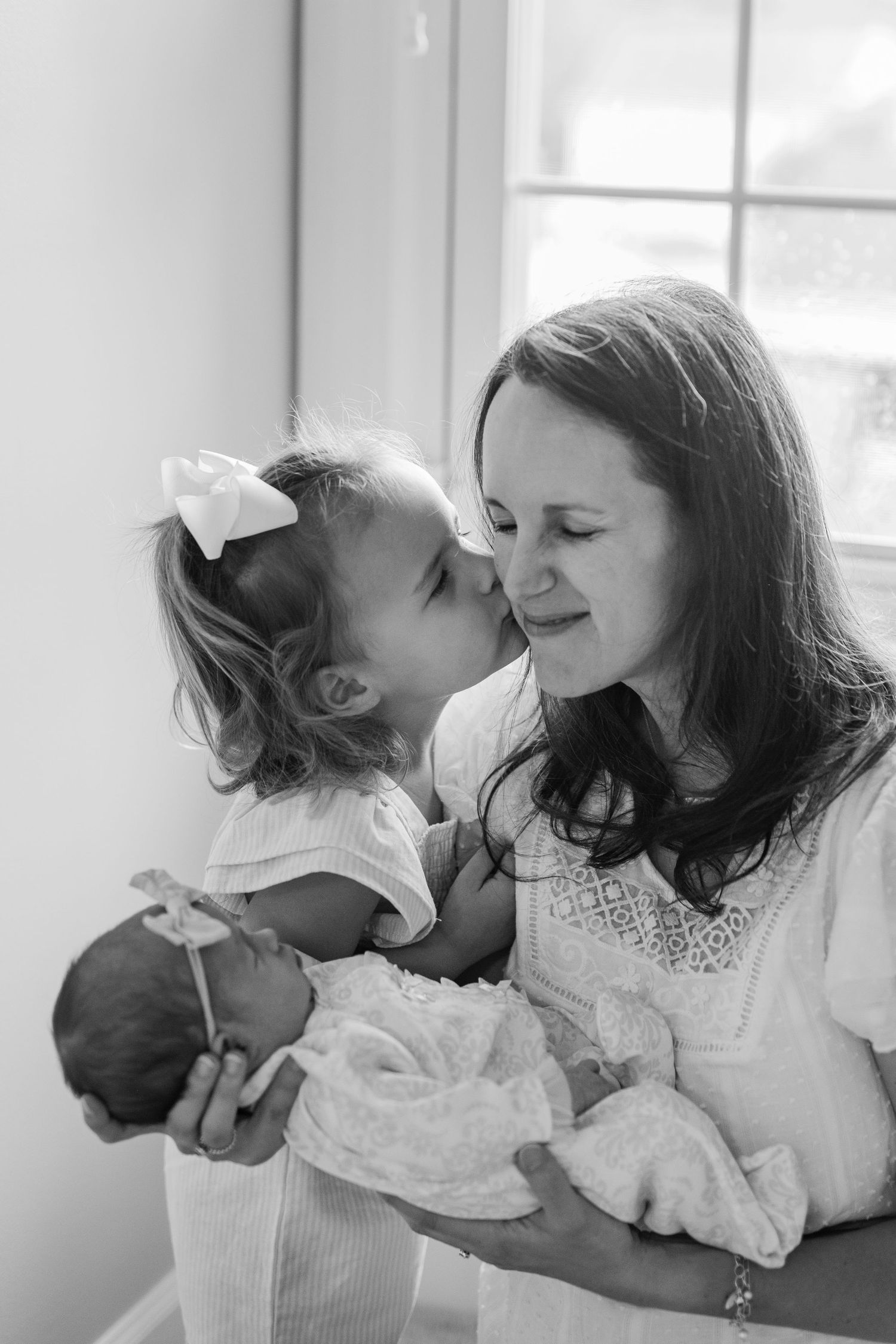 mom toddler and baby. toddler kissing mother's cheek. black and white photo. at home newborn session.