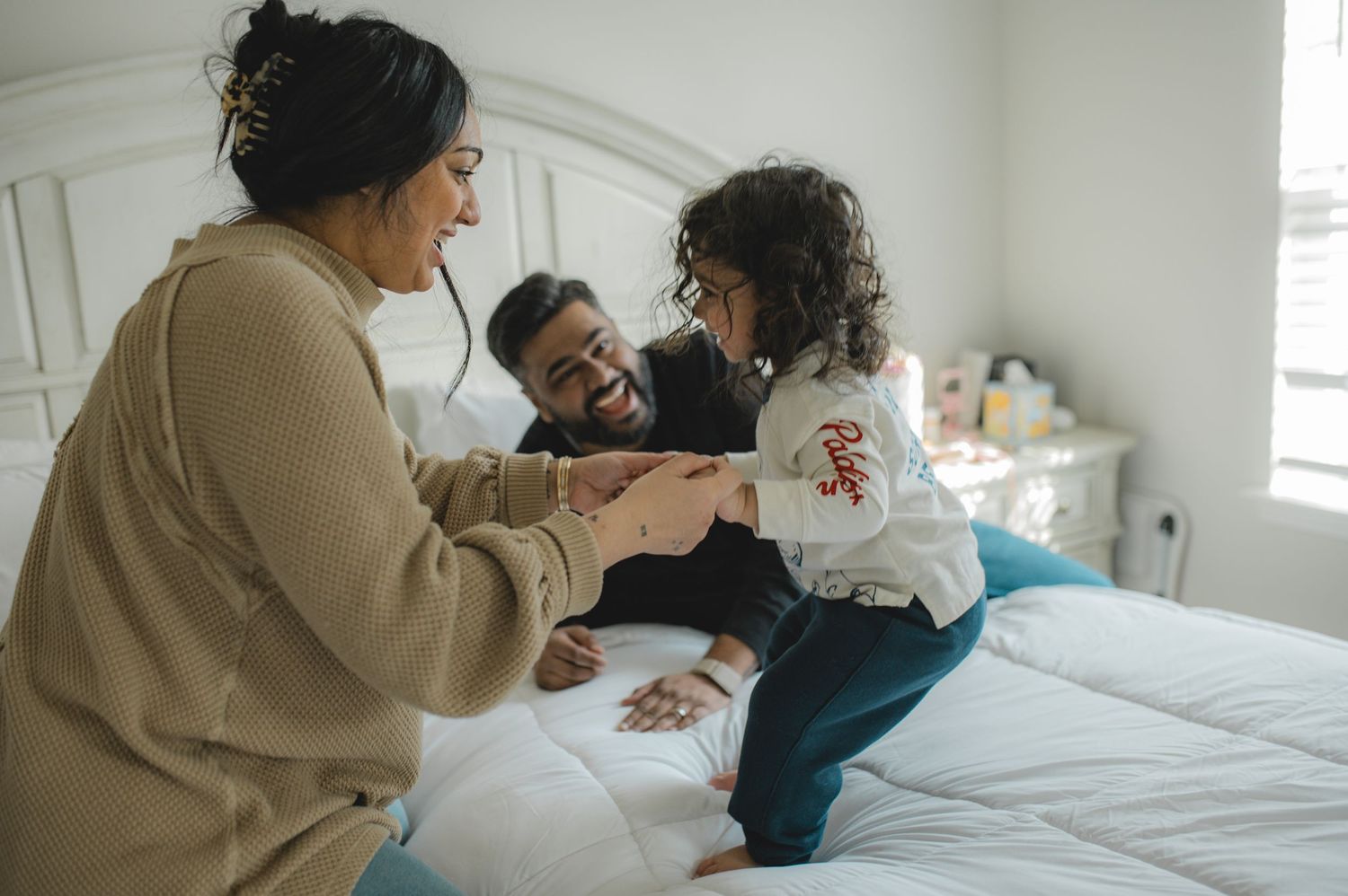 mom dad and son jumping on bed, holding hands, playing, laughing, smiling
