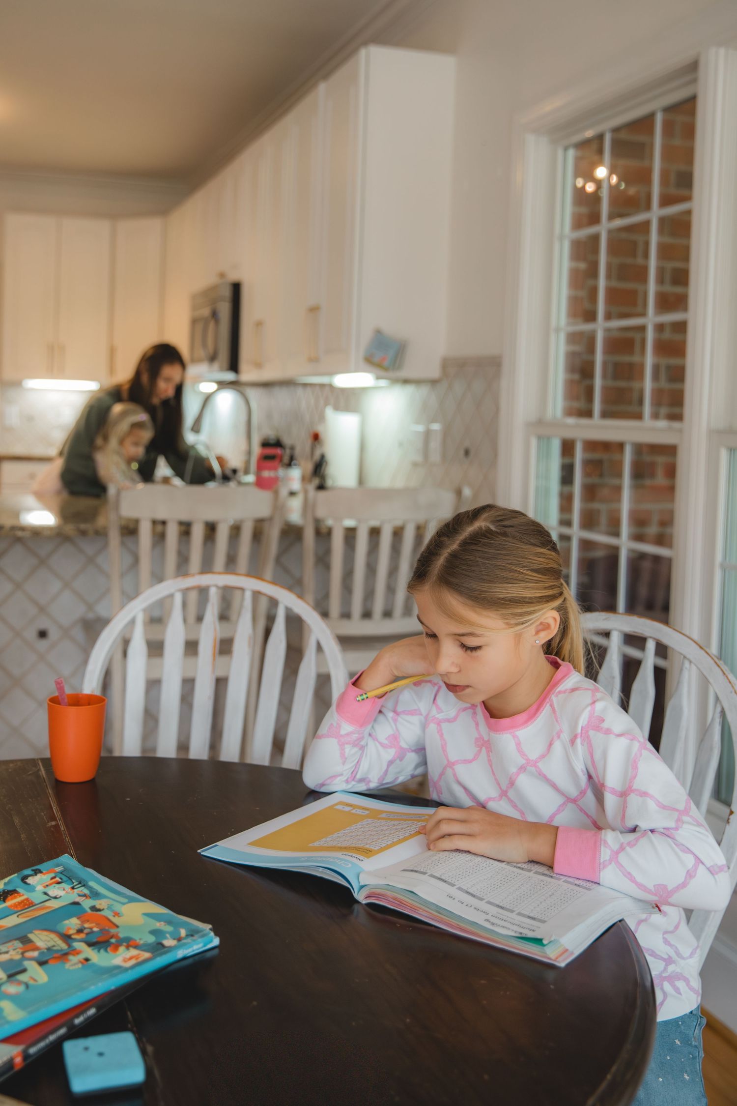 child sitting at kitchen table doing school work, homeschooling, mom and child in background washing hands in kitchen sink
