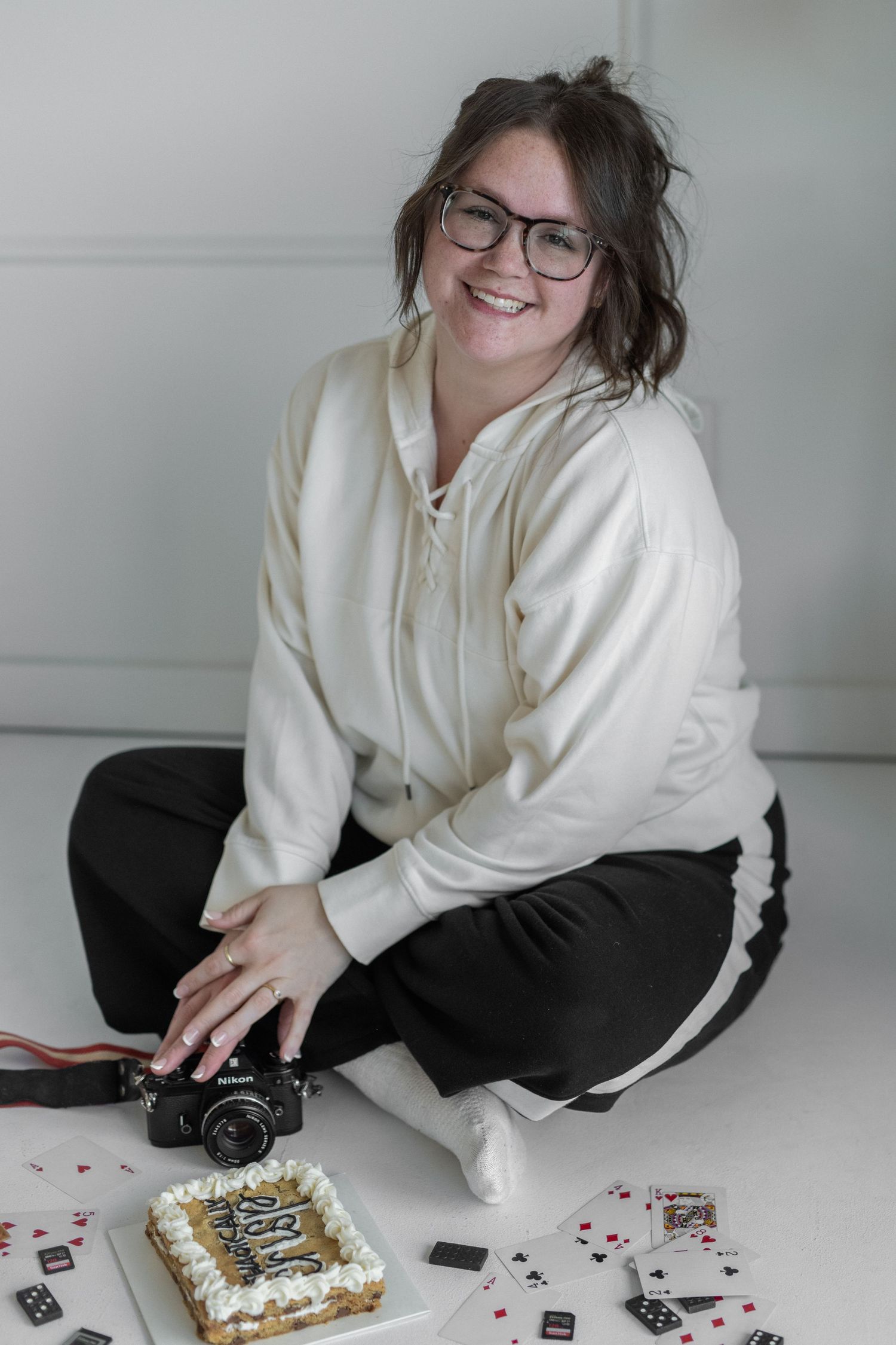 woman smiling at the camera. sweatsuit outfit. long brown hair in ponytail. camera and cookie cake on floor. domino set, playing cards