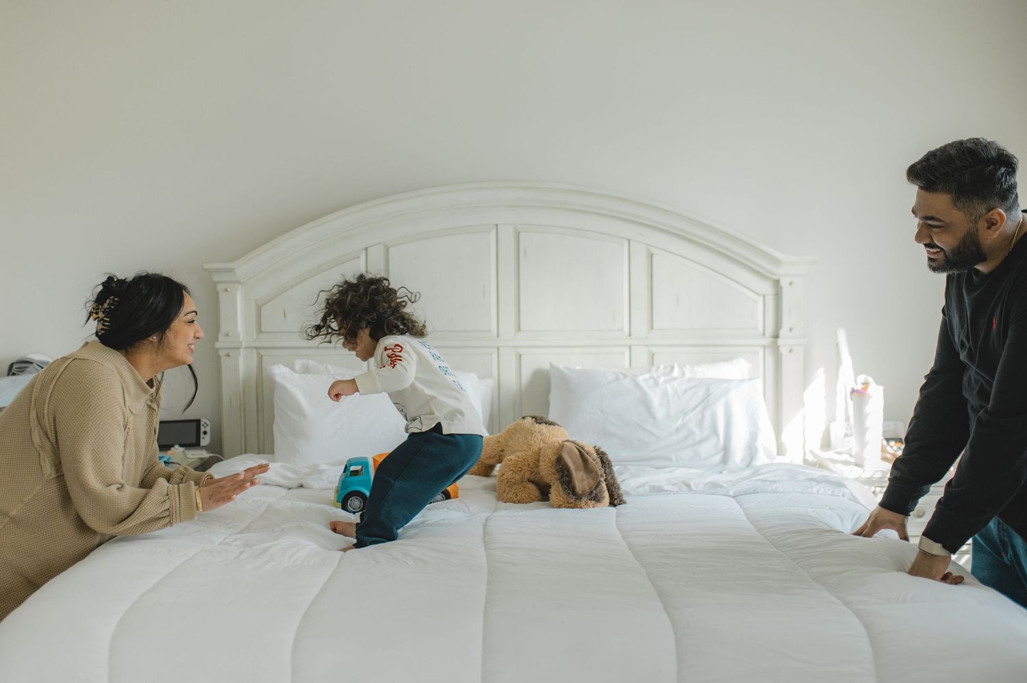mom dad and son jumping on bed, holding hands, playing, laughing, smiling