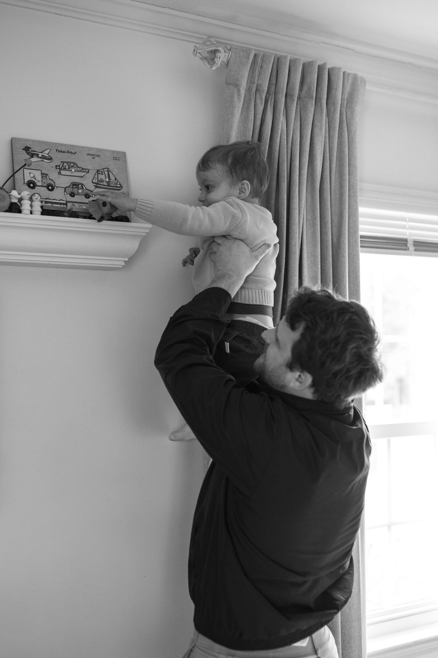 dad holding child up to get a wooden toy off of shelf in front of window