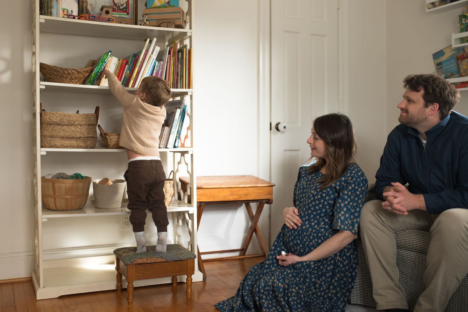 mom and dad sitting. toddler reaching for book on bookshelf standing on a stool. pregnant mama holding belly smiling. at home family session