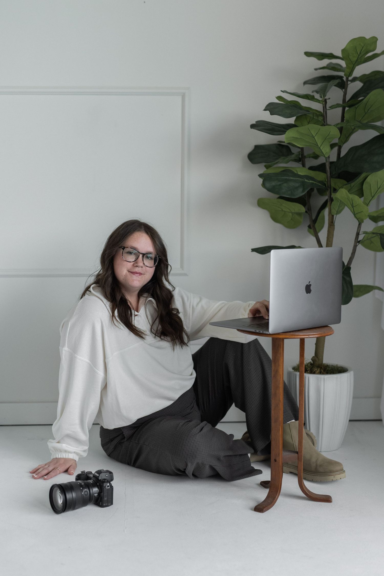 woman smiling at the camera. business casual outfit. long brown hair. fiddle-leaf fig tree in the background. laptop on table. camera