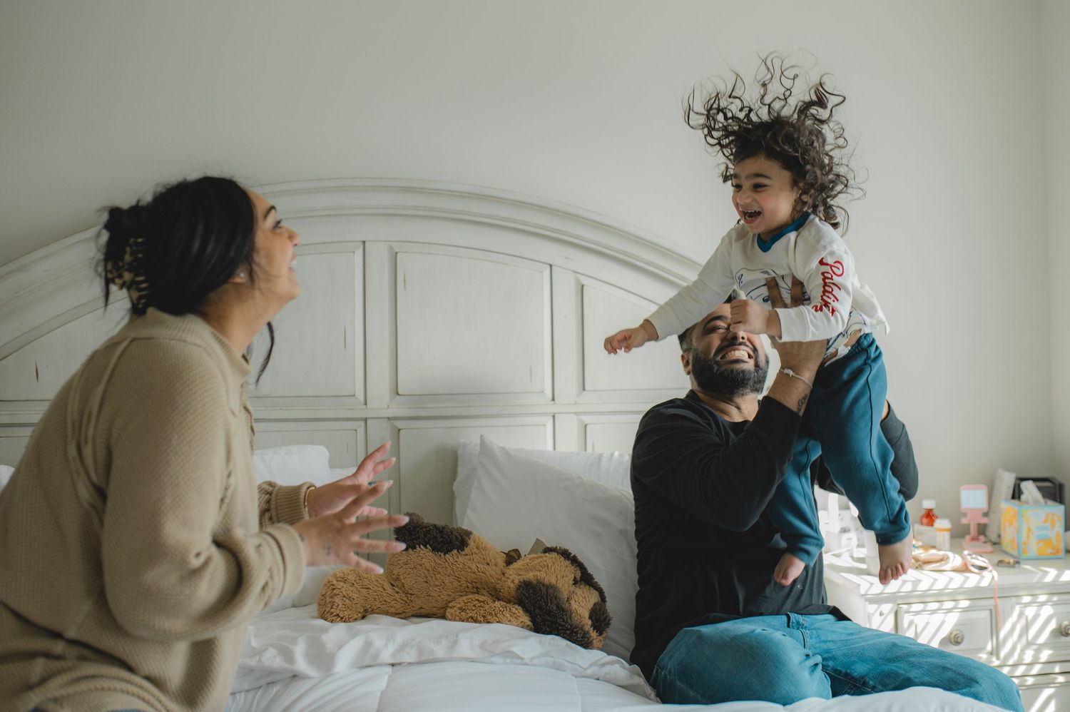 mom dad and son jumping on bed, holding hands, playing, laughing, smiling
