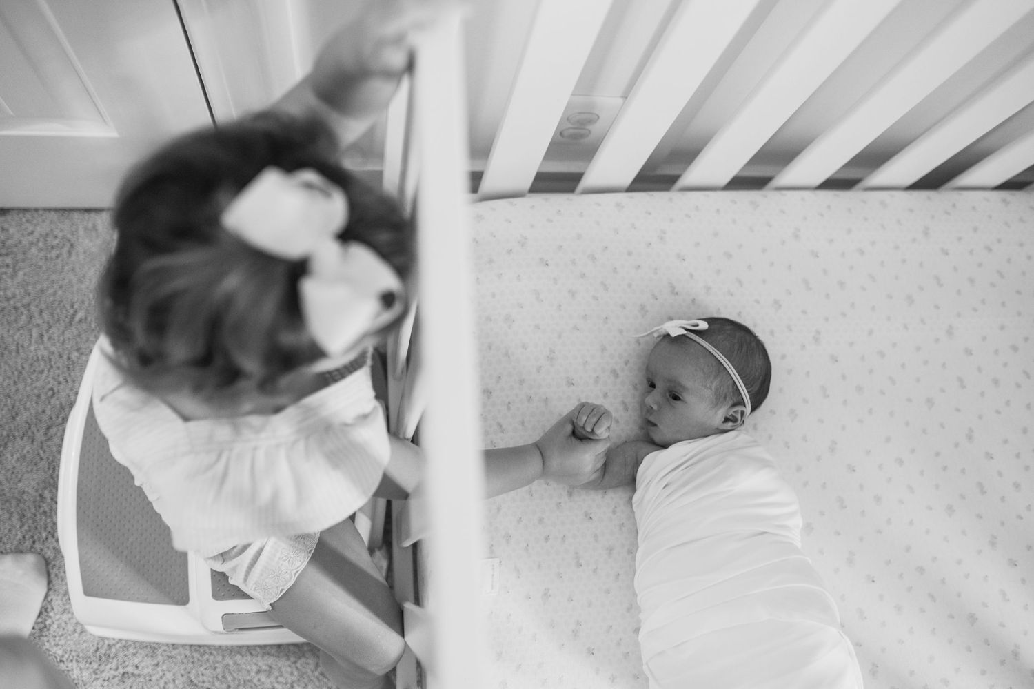black and white photo. toddler girl holding newborn baby hand through crib slats. at home documentary newborn session.