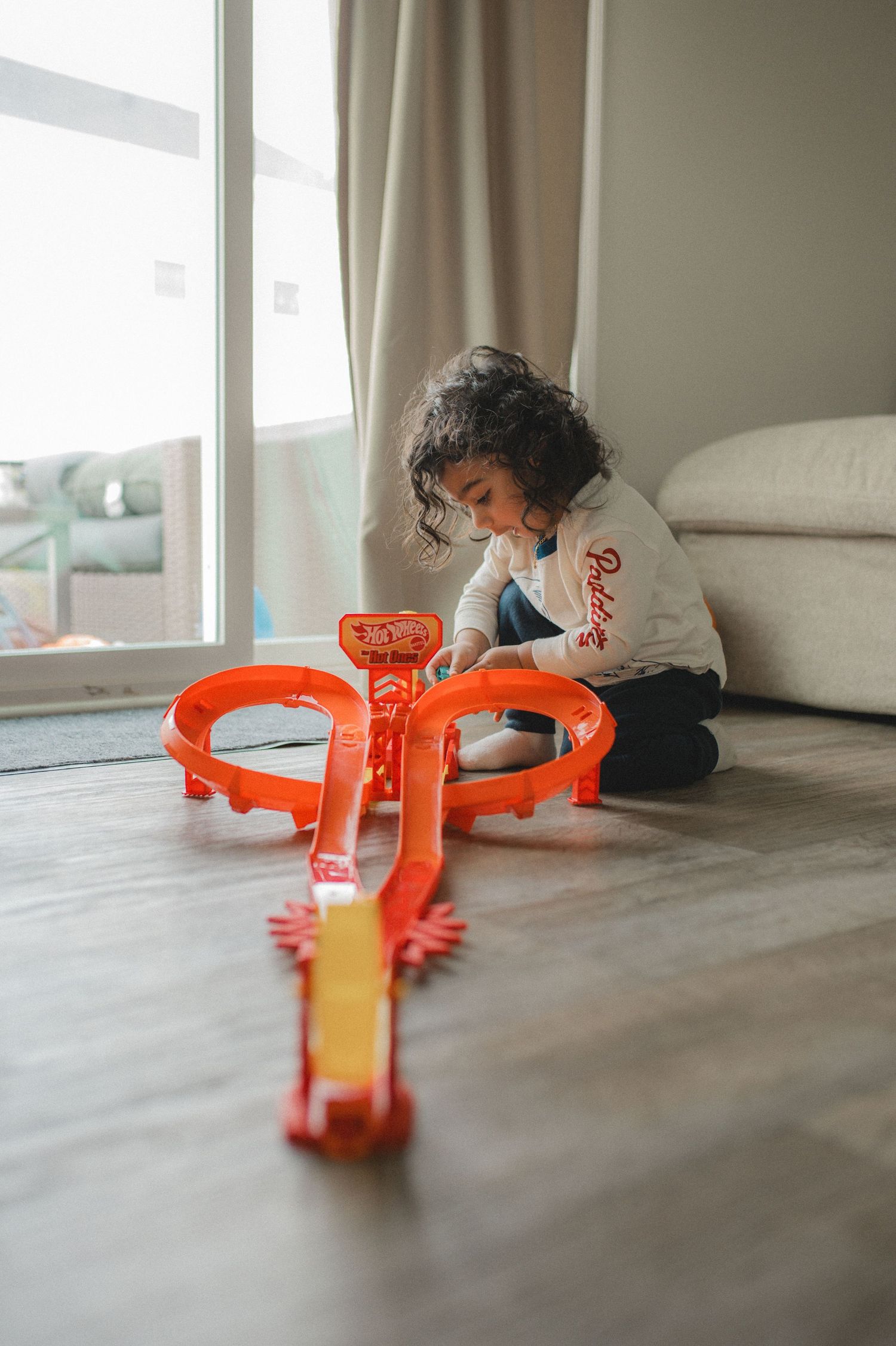 child playing with hot wheels car race track on floor in front of glass door window