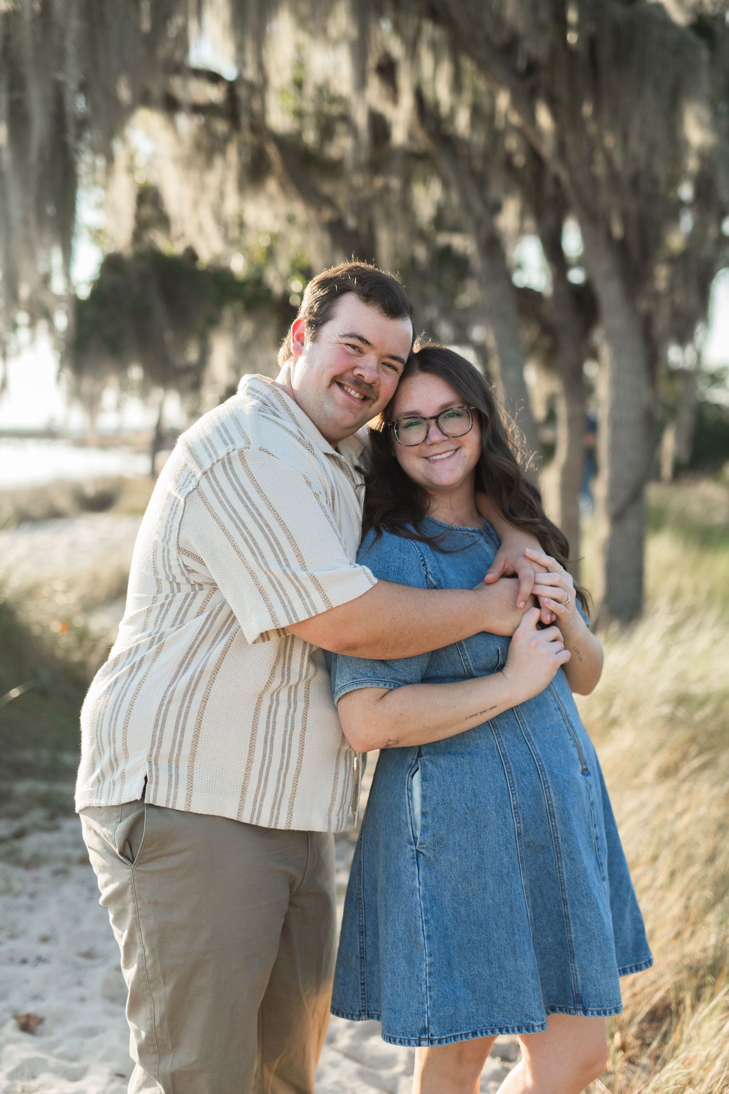 husband and wife. hugging. smiling. beach. spanish moss. sand. trees. grass.