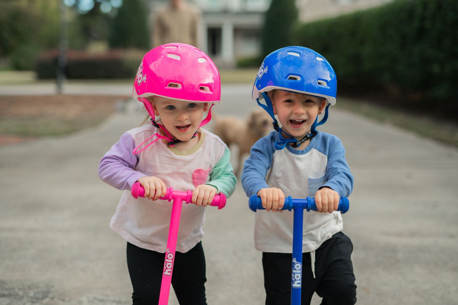 twins. boy and girl. riding scooters in driveway. wearing helmets. pink and blue. laughing, having fun. at home documentary family session