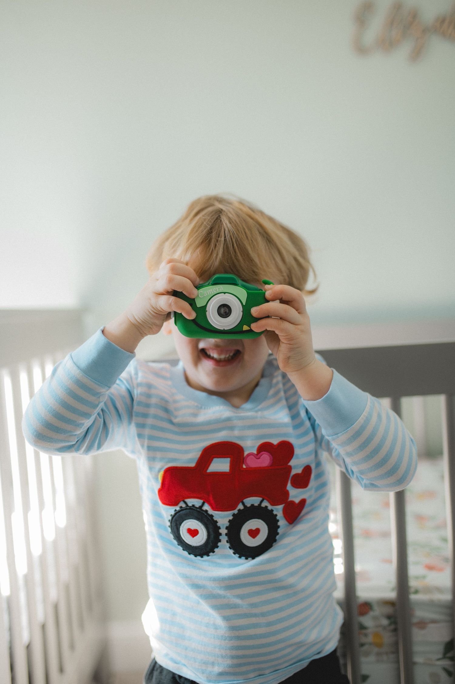 child holding camera in front of crib in nursery with red truck on shirt