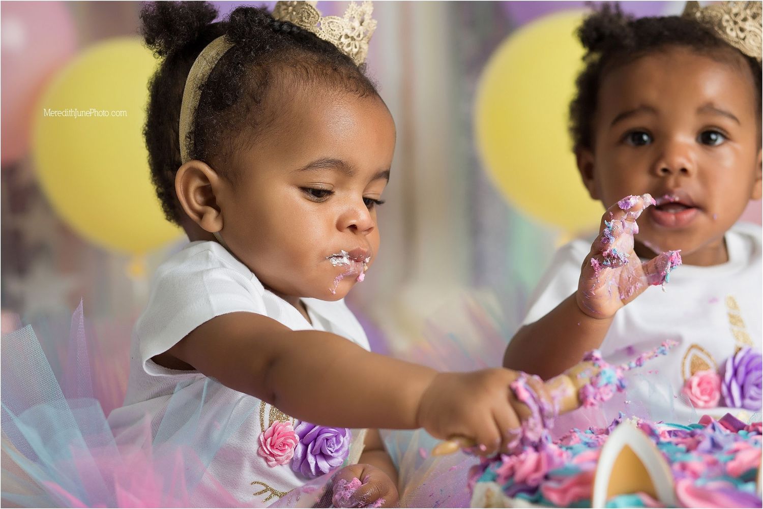 Two toddlers in pink tutus and party crowns celebrate with colorful balloons and cake at a birthday party.