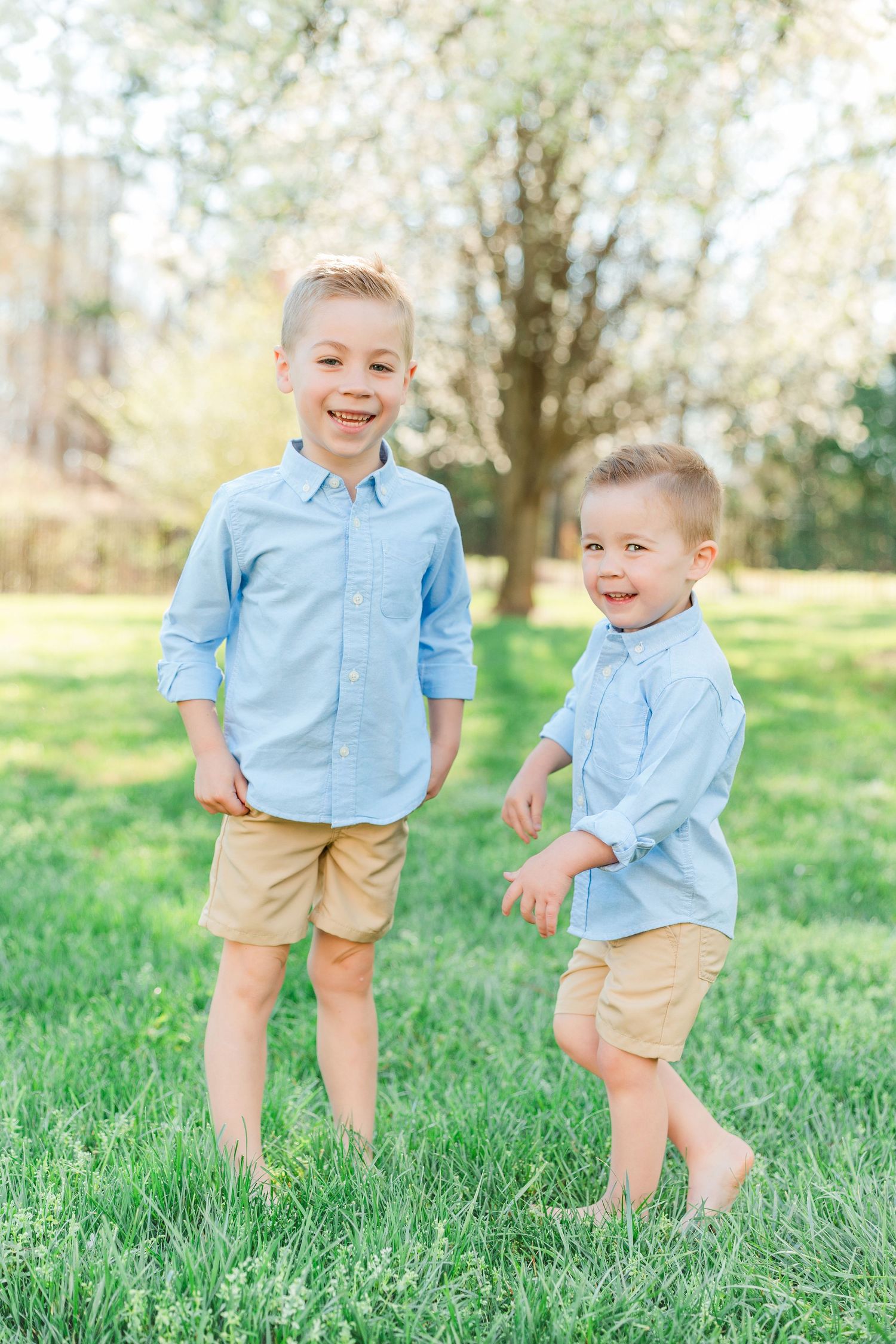 Two children wearing matching light blue shirts and khaki shorts laugh together on green grass in a sunny outdoor setting.