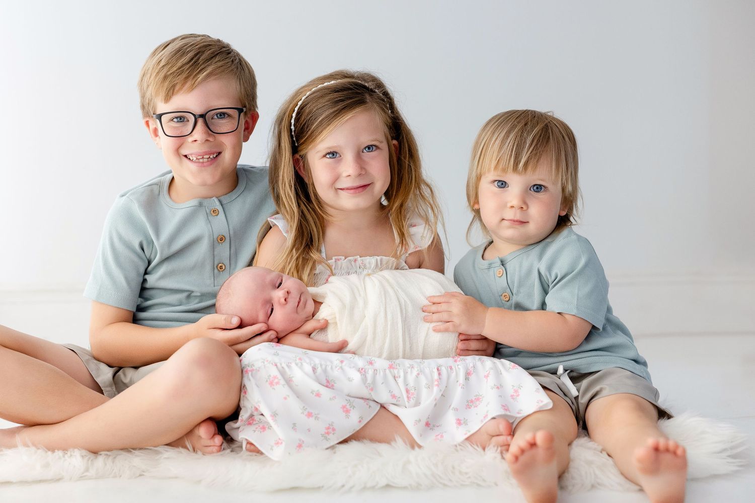 Three siblings pose together on white backdrop, older boy in glasses holding newborn baby wrapped in white.