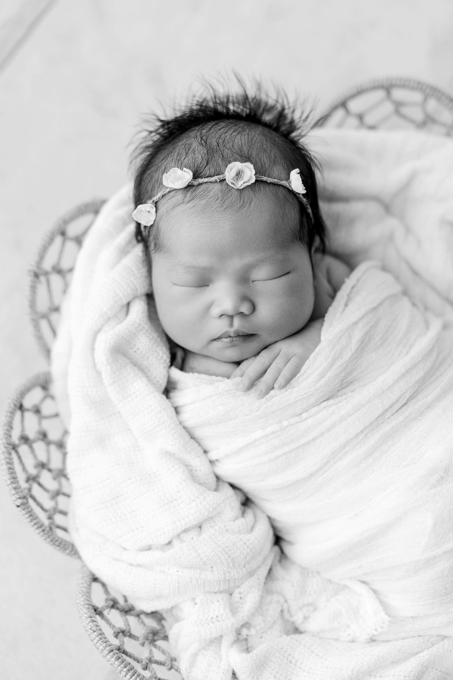 Black and white photo of a sleeping newborn baby girl wrapped in white swaddle wearing a delicate floral headband.