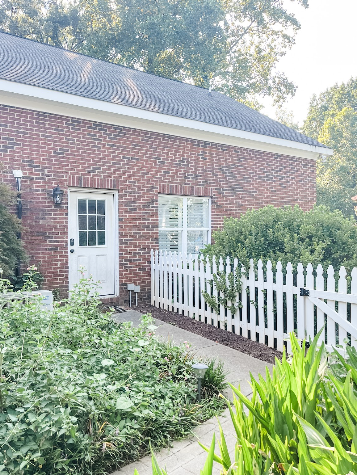 A red brick ranch house with white picket fence and landscaped garden featuring lush greenery and ornamental plants.