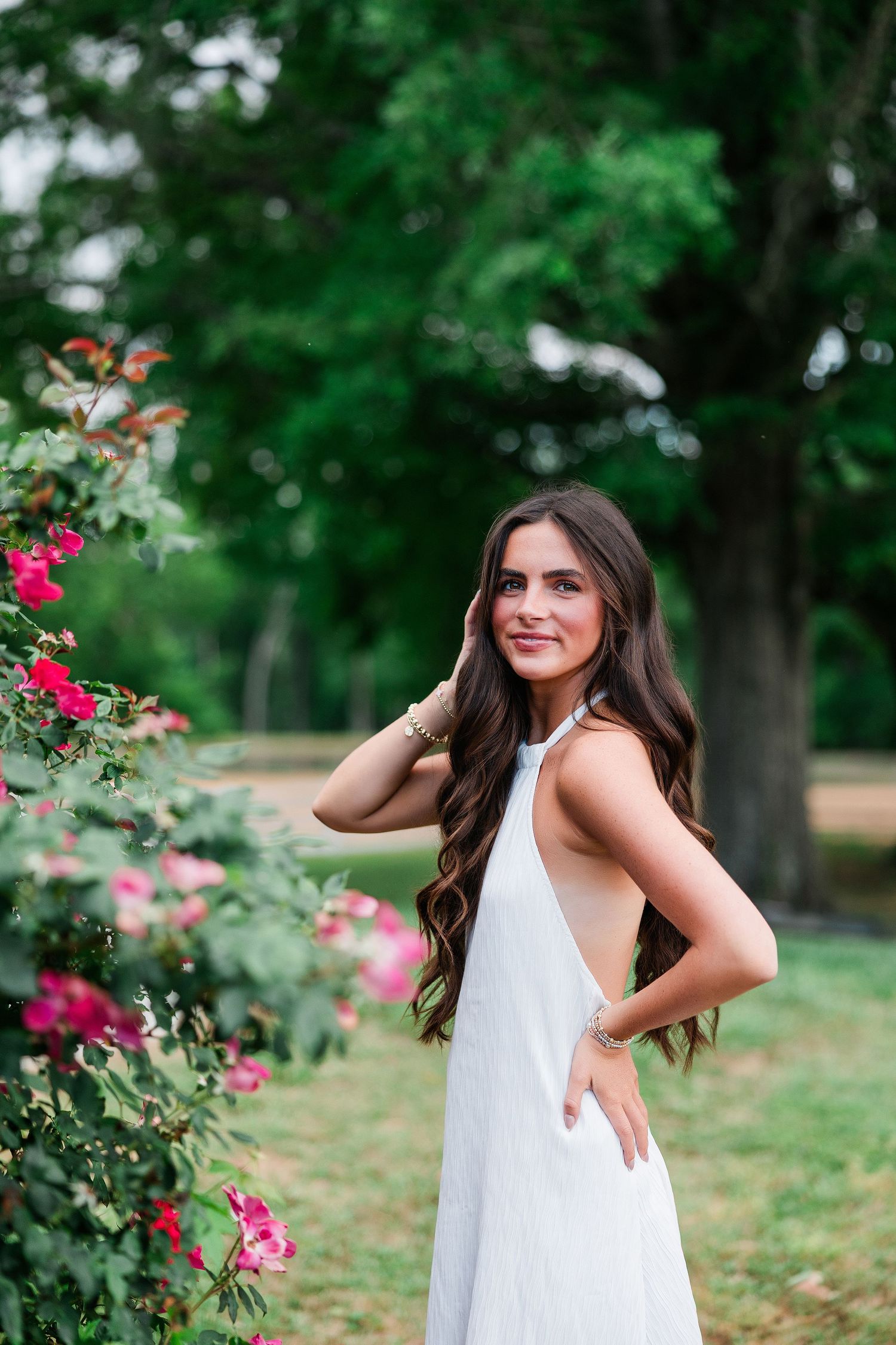 Young woman in white halter dress poses gracefully beside pink roses in a lush green park setting.