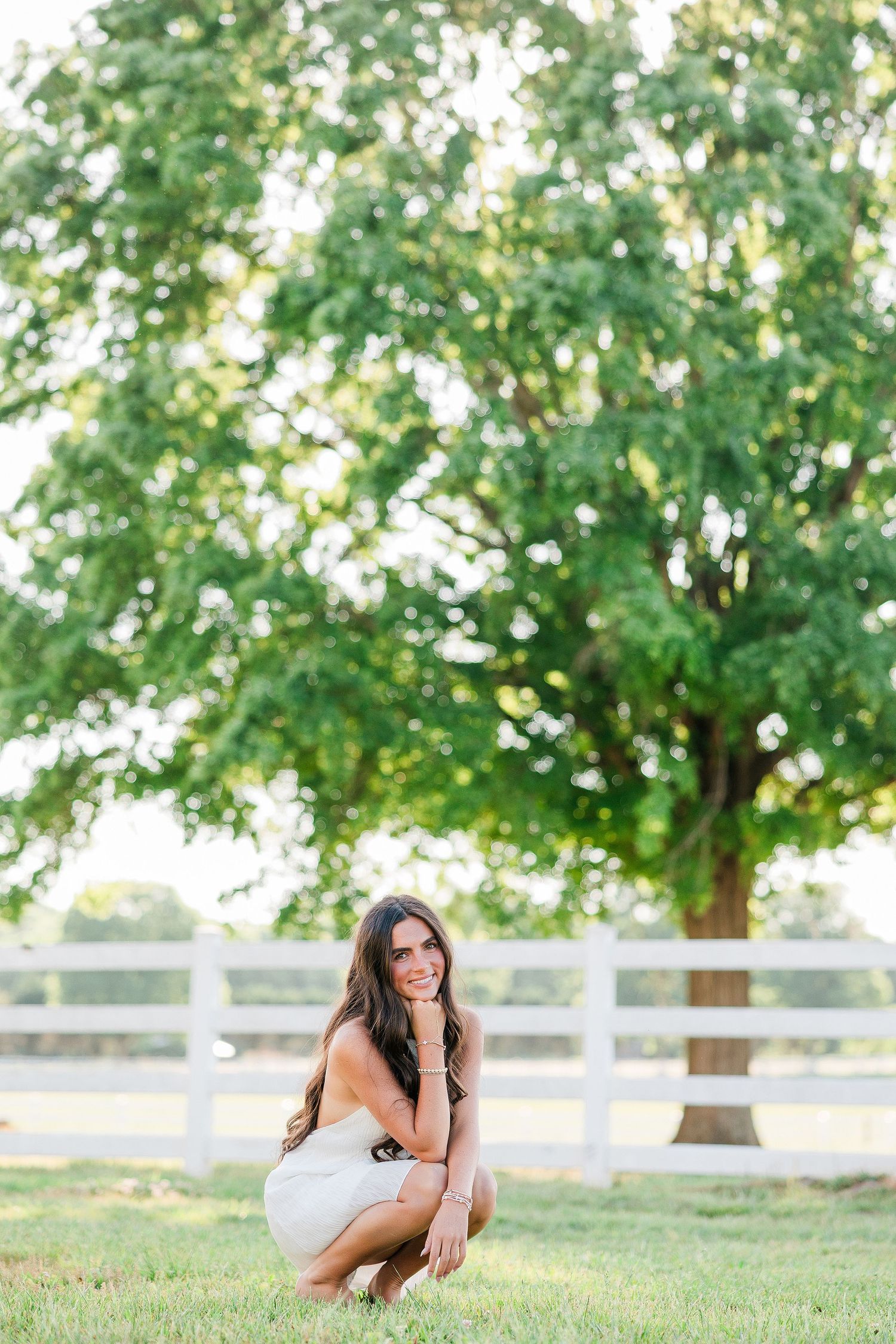 Woman in white dress crouching on green grass near white fence and large tree outdoors.
