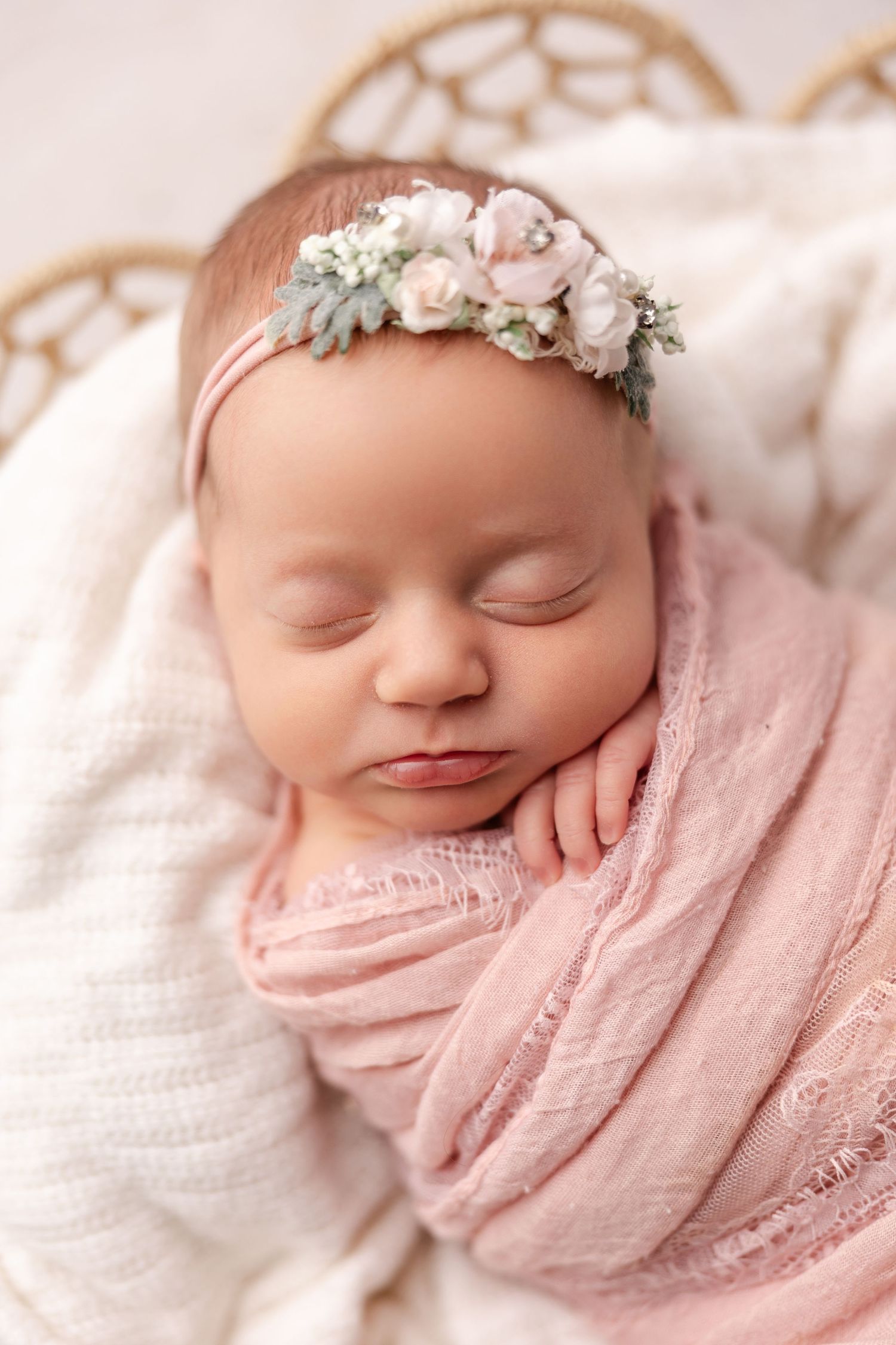 Sleeping newborn wrapped in soft pink swaddle wearing delicate floral headband on white textured backdrop.