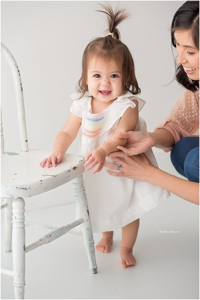 A small child in a white dress stands beside a vintage white chair while being supported by caring hands.