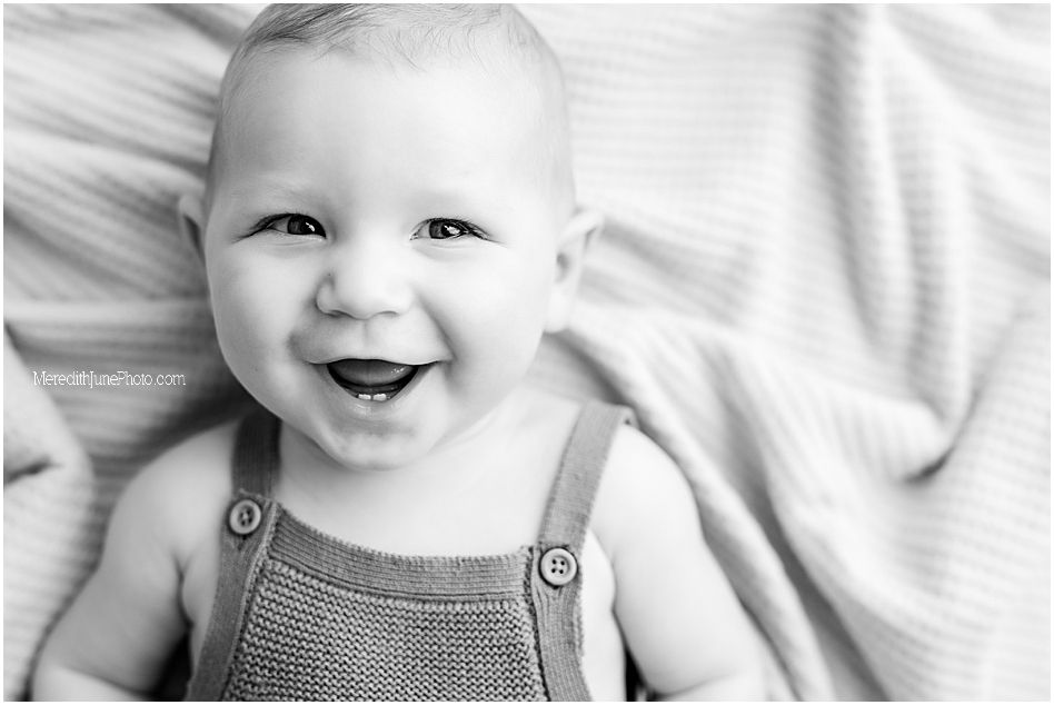 Black and white photo series showing a smiling baby wearing overalls while lying on white bedding.