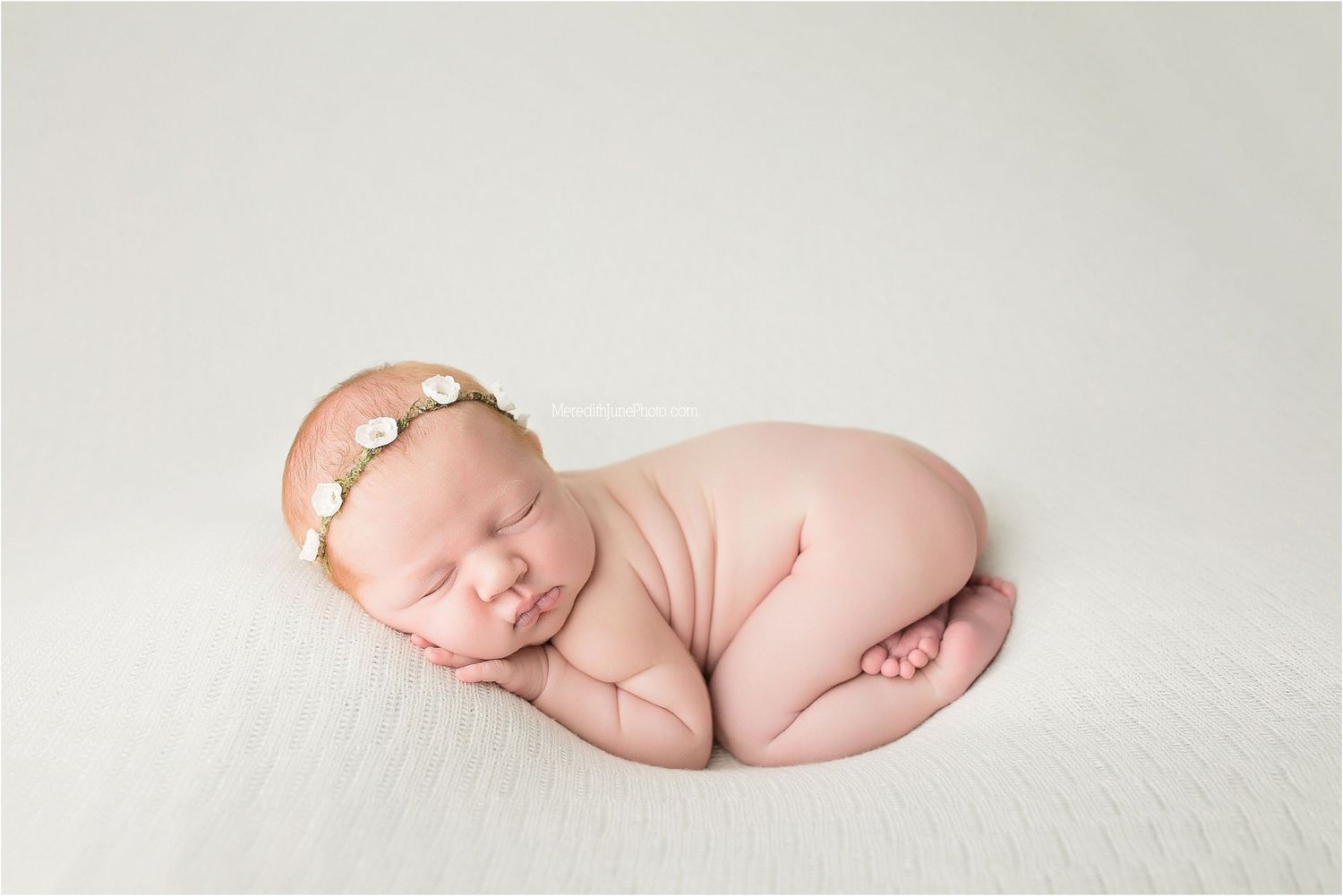 Peaceful sleeping newborn curled up on soft white blanket wearing delicate peach headband.