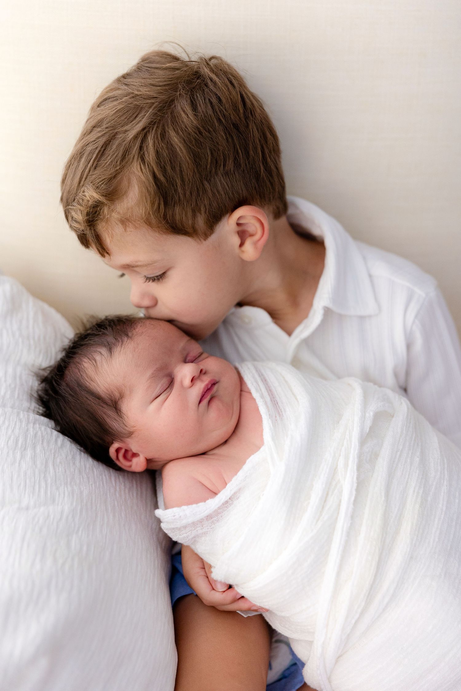 Older boy in white shirt kissing sleeping newborn baby swaddled in white muslin wrap.
