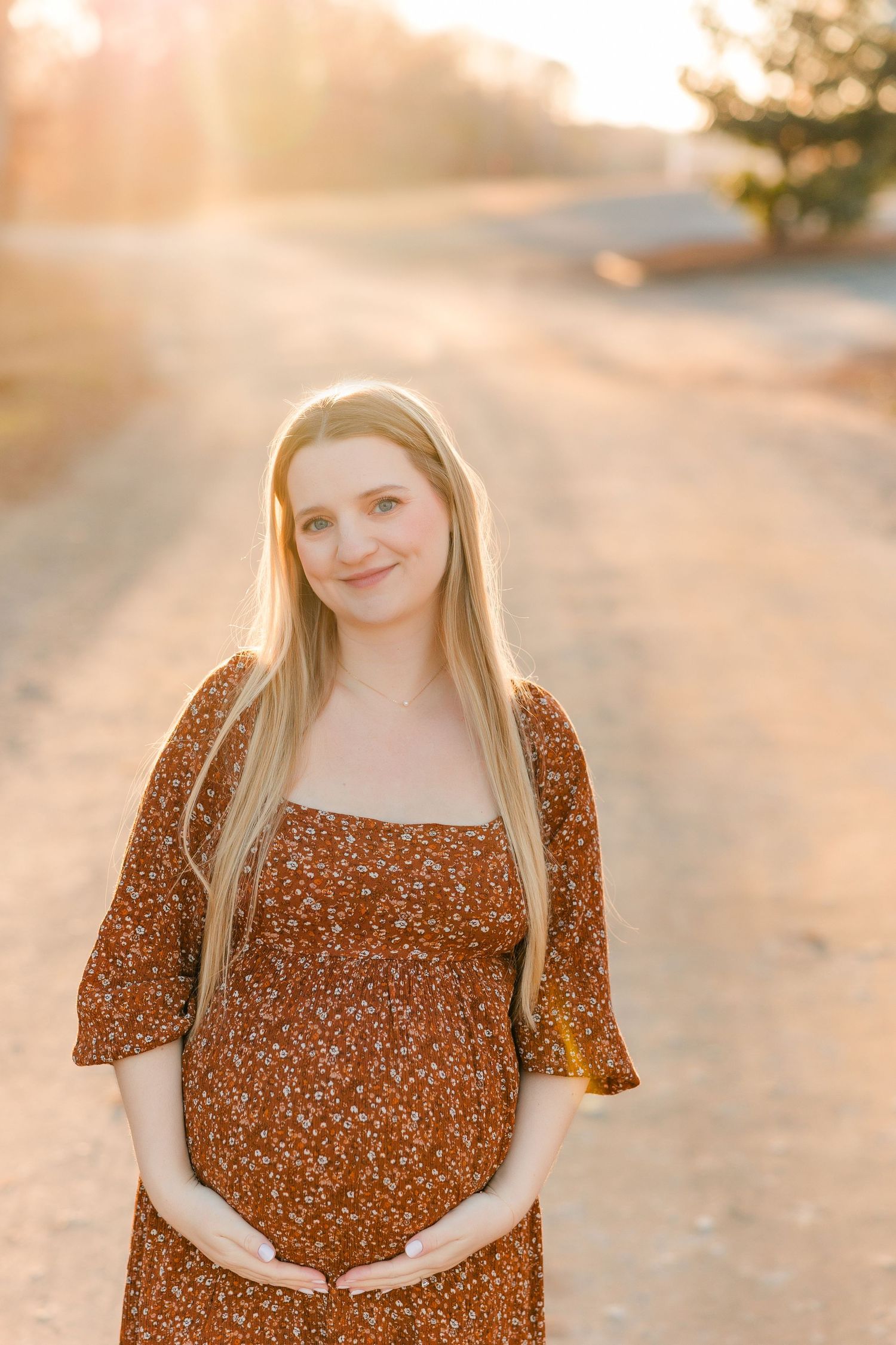A maternity photoshoot in warm sunset lighting featuring a brown floral dress against a natural outdoor backdrop.